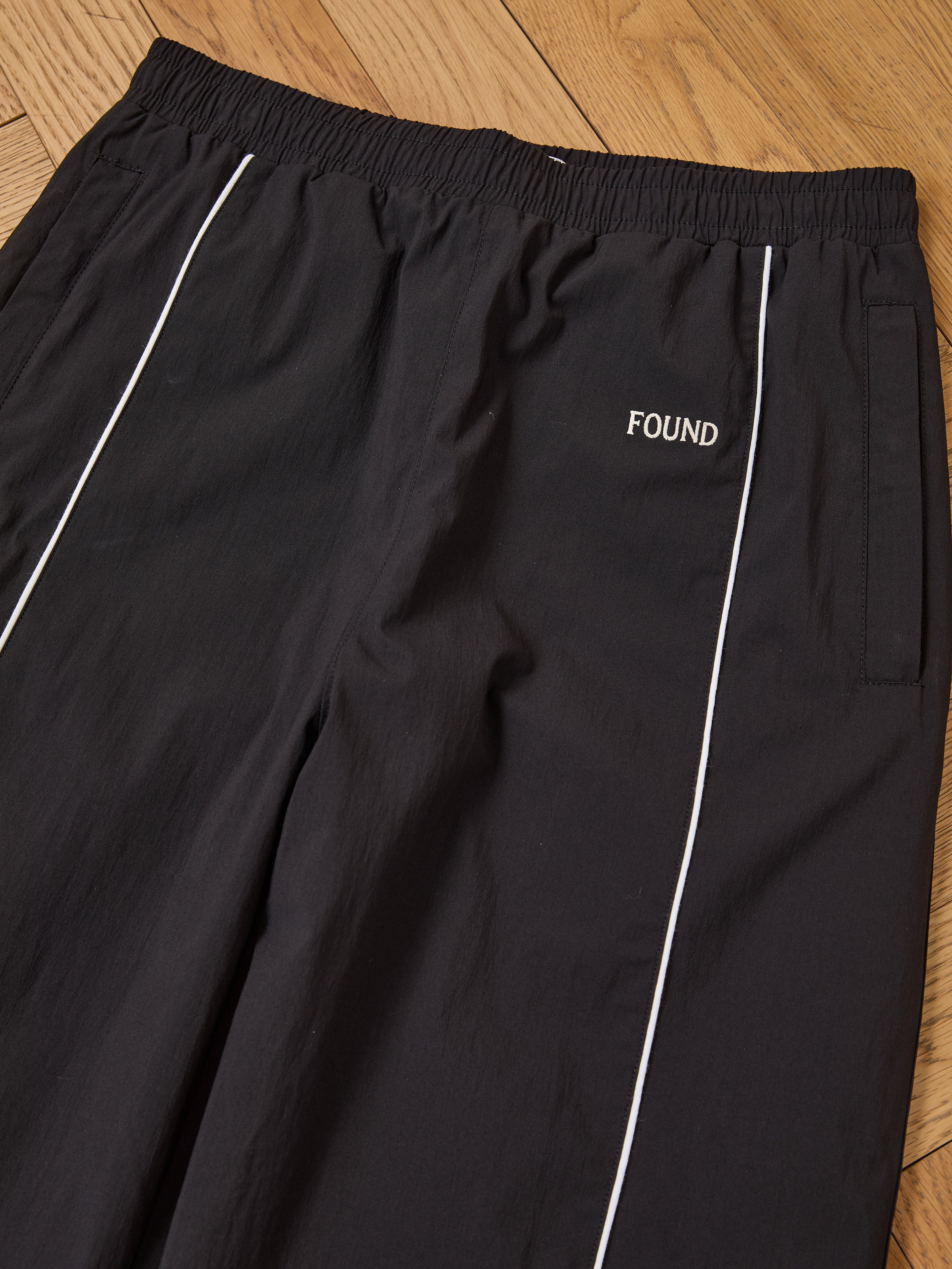 The Found Arena Stripe Track Pants feature white piping, an adjustable waistband, side pockets, and "FOUND" embroidery near the left pocket. Laid on a wooden floor, these unisex pants blend comfort and style for any activity.