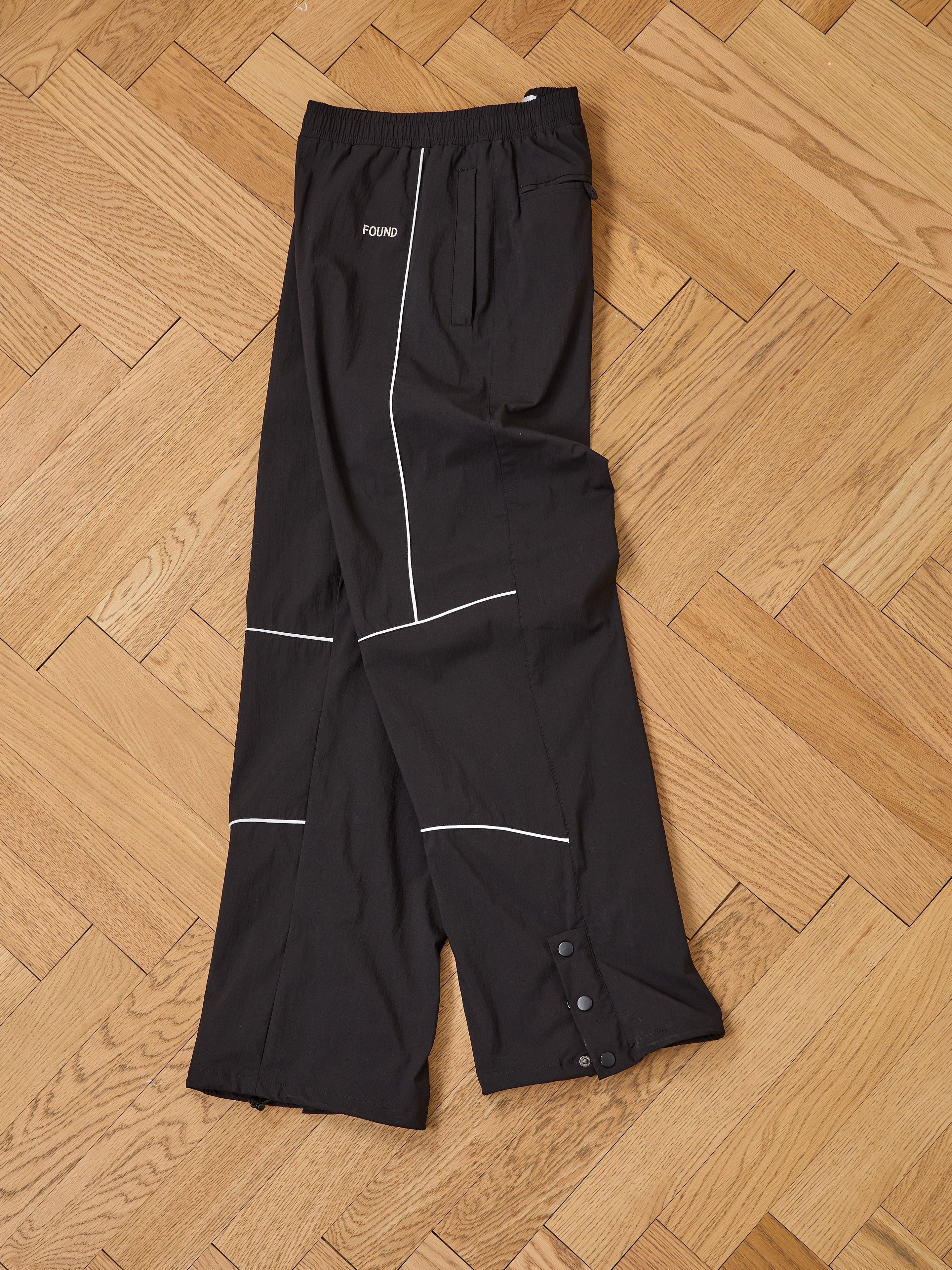 The Found Arena Stripe Track Pants, featuring white piping, adjustable waistband, side pockets, and buttoned cuffs, are laid flat on a herringbone floor—perfect unisex sportswear for everyday comfort.