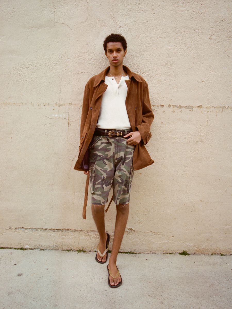 A person stands against a beige wall wearing a brown jacket, white shirt, and FOUND Camouflage Twill Utility Cargo Shorts in a Unisex Relaxed Fit, styled with a black belt and flip-flops for a subtle vintage vibe.