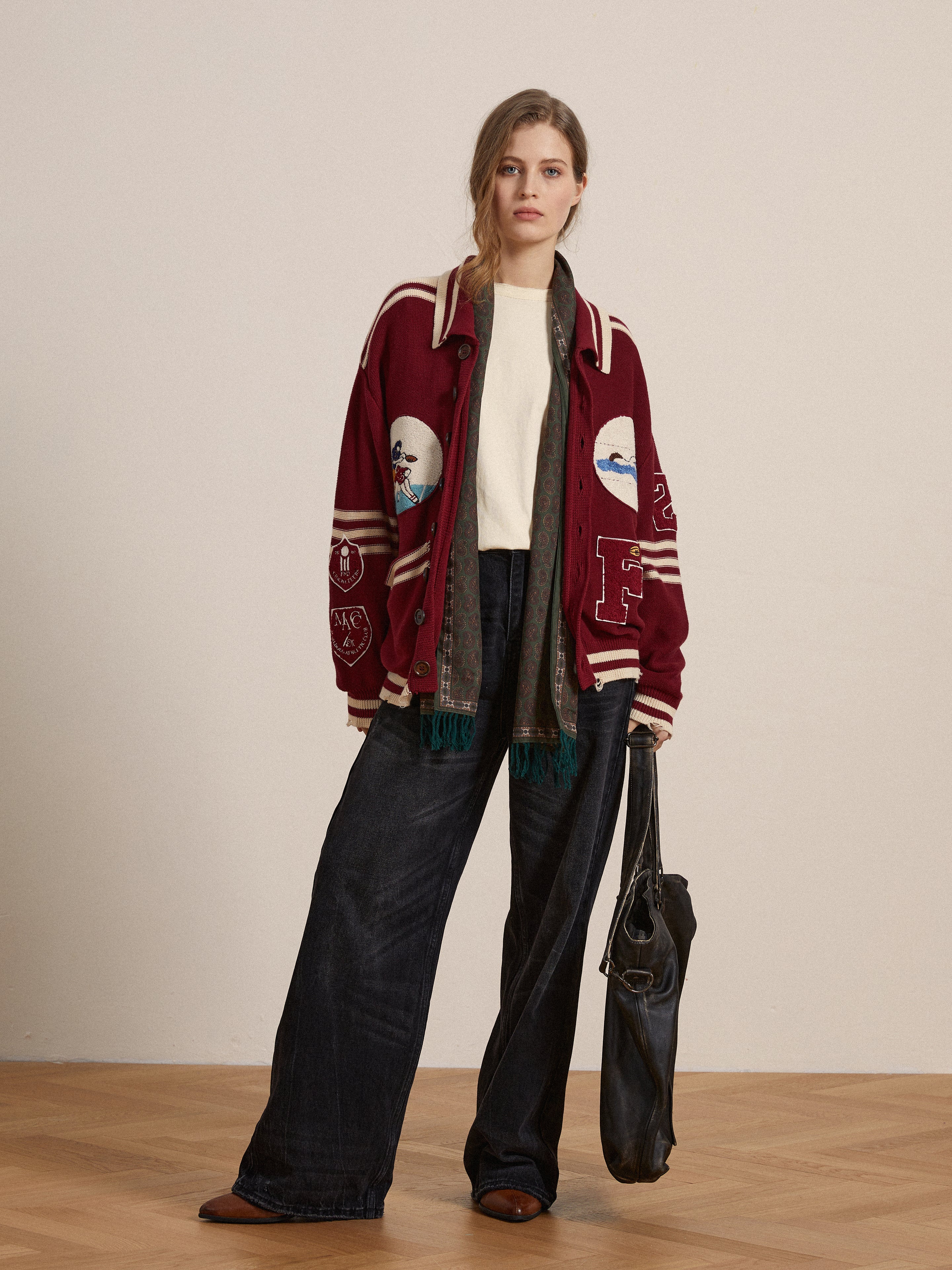 A woman stands indoors wearing the FOUND Reversed LS Tee—a white long-sleeve with a distressed neckline—layered under a maroon varsity jacket, paired with wide-leg dark jeans, brown shoes, and holding a large black bag.