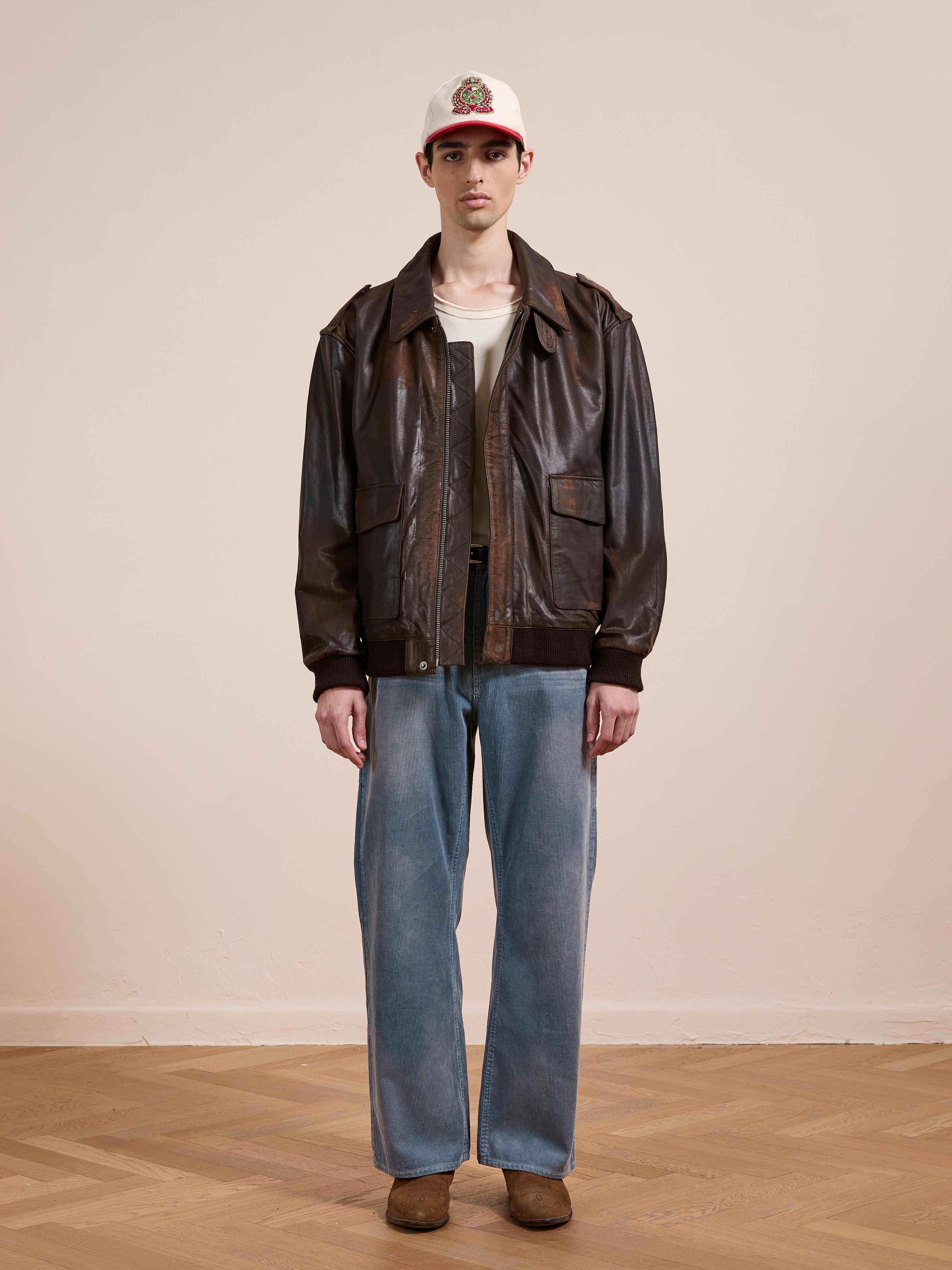 A person stands indoors against a beige wall and wooden floor, wearing the FOUND Distressed Leather Flight Jacket over a white shirt, with a white cap, loose blue jeans, and brown shoes.