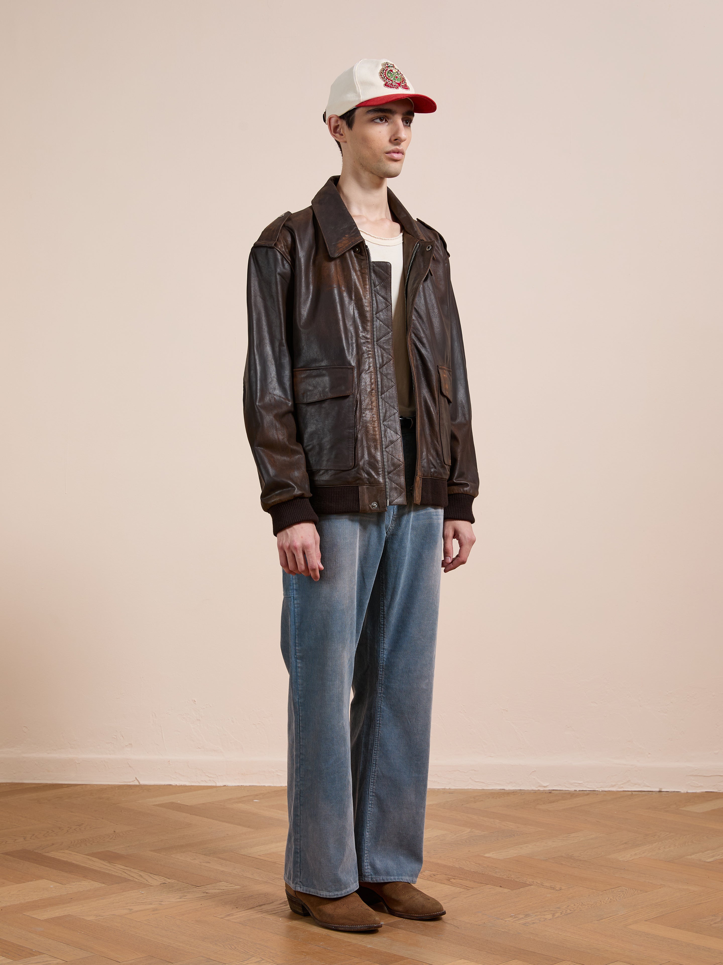 Indoors, a person wears the FOUND Distressed Leather Flight Jacket, a white and red cap, white t-shirt, wide-leg blue jeans, and brown boots on a wooden floor.