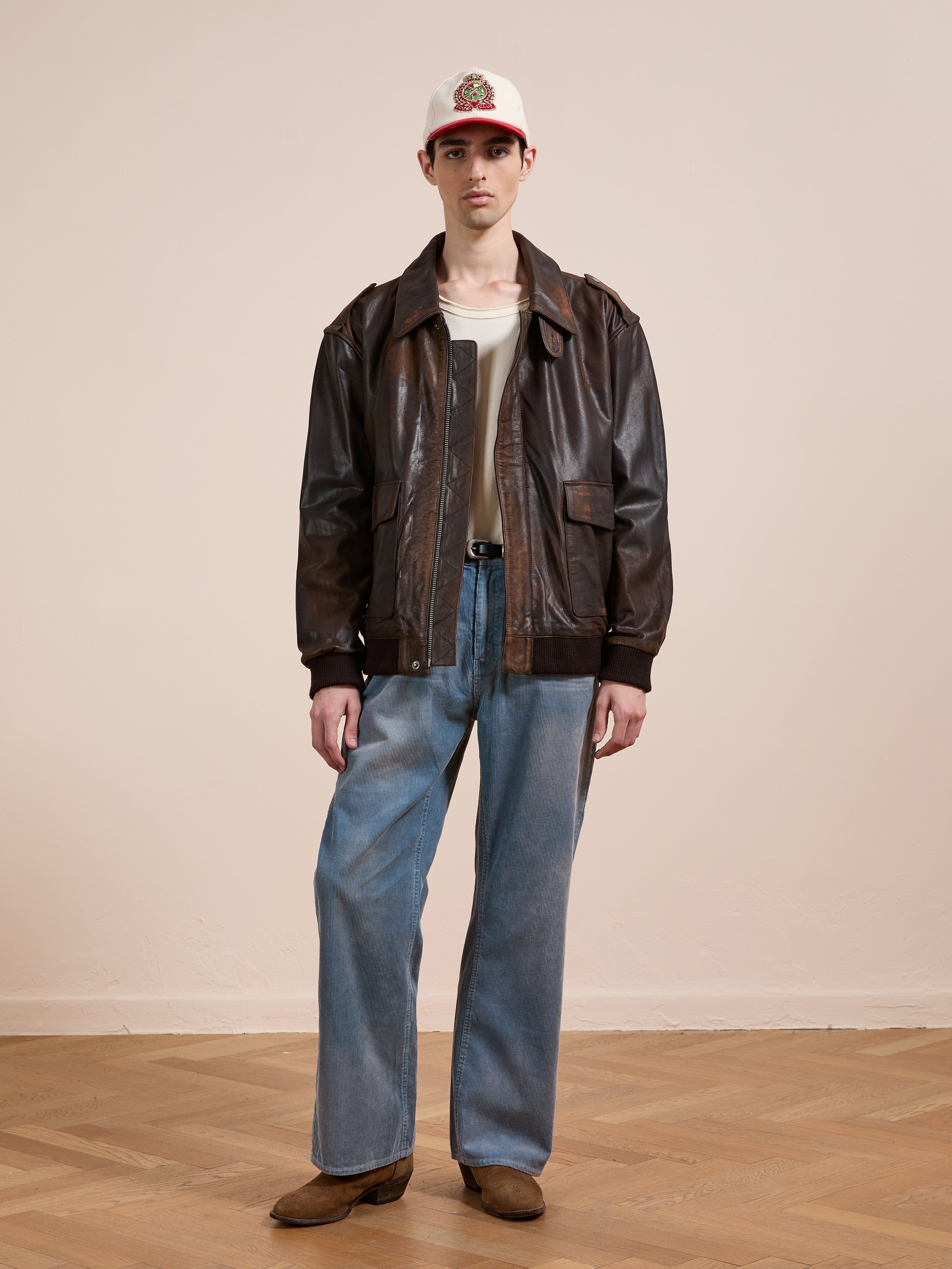 Indoors, a person wears the FOUND Distressed Leather Flight Jacket over a light t-shirt, wide blue jeans, brown shoes, and a white baseball cap with a patch.