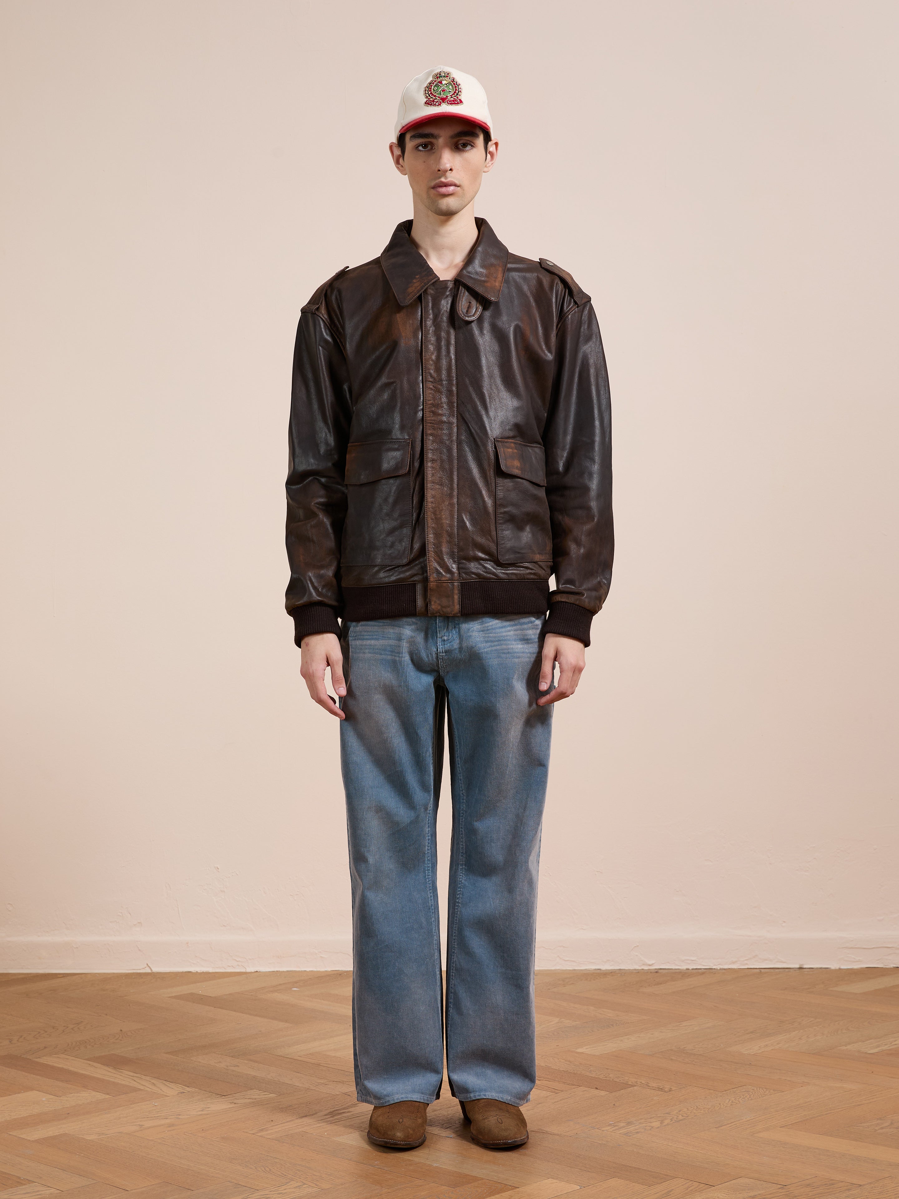 Indoors against a plain wall, a person wears the FOUND Distressed Leather Flight Jacket, paired with blue jeans, brown shoes, and a white baseball cap.