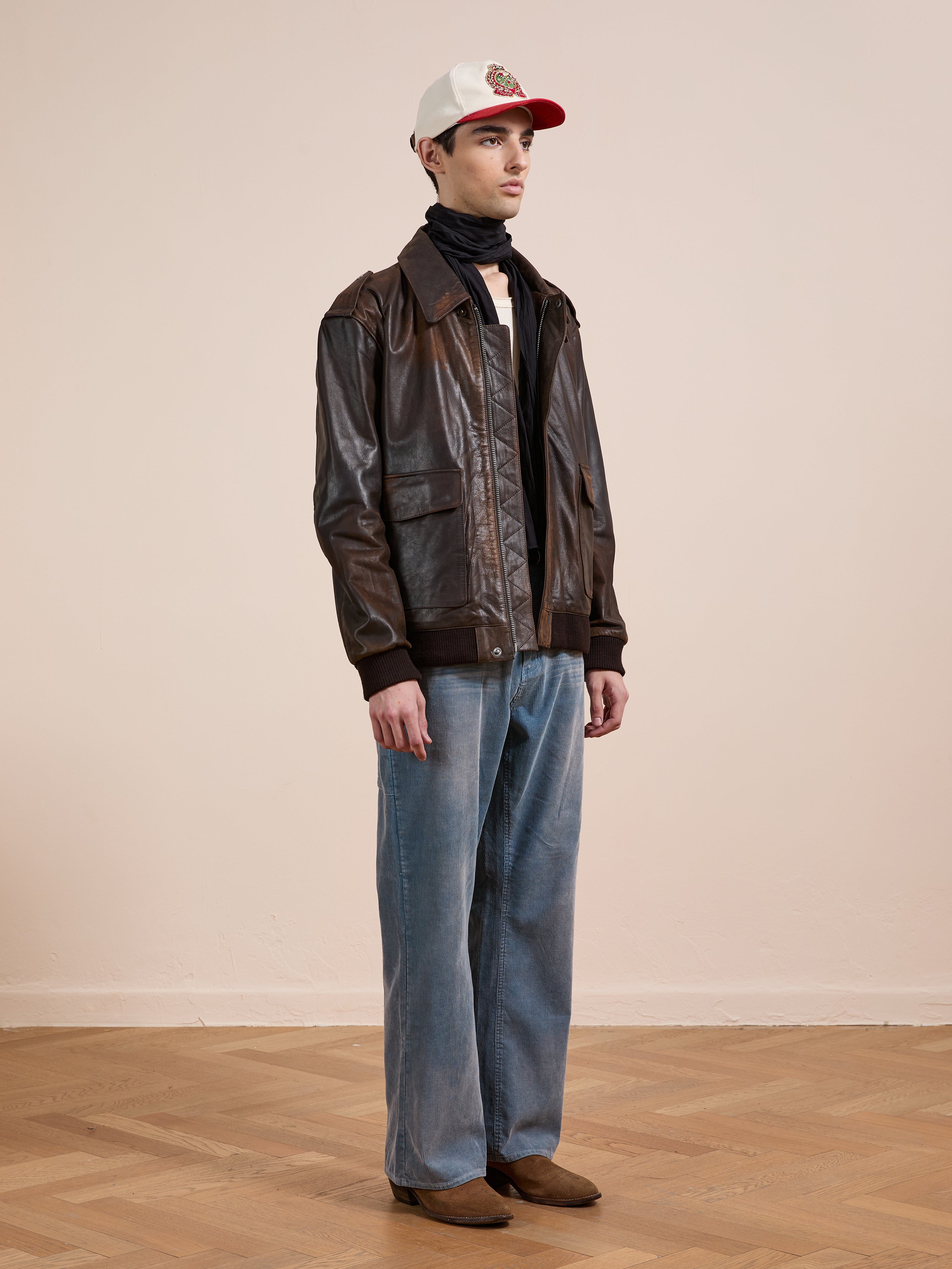 A person in a white cap, black scarf, FOUND Distressed Leather Flight Jacket, loose blue jeans, and brown shoes stands on a wooden floor before a plain light-colored wall.