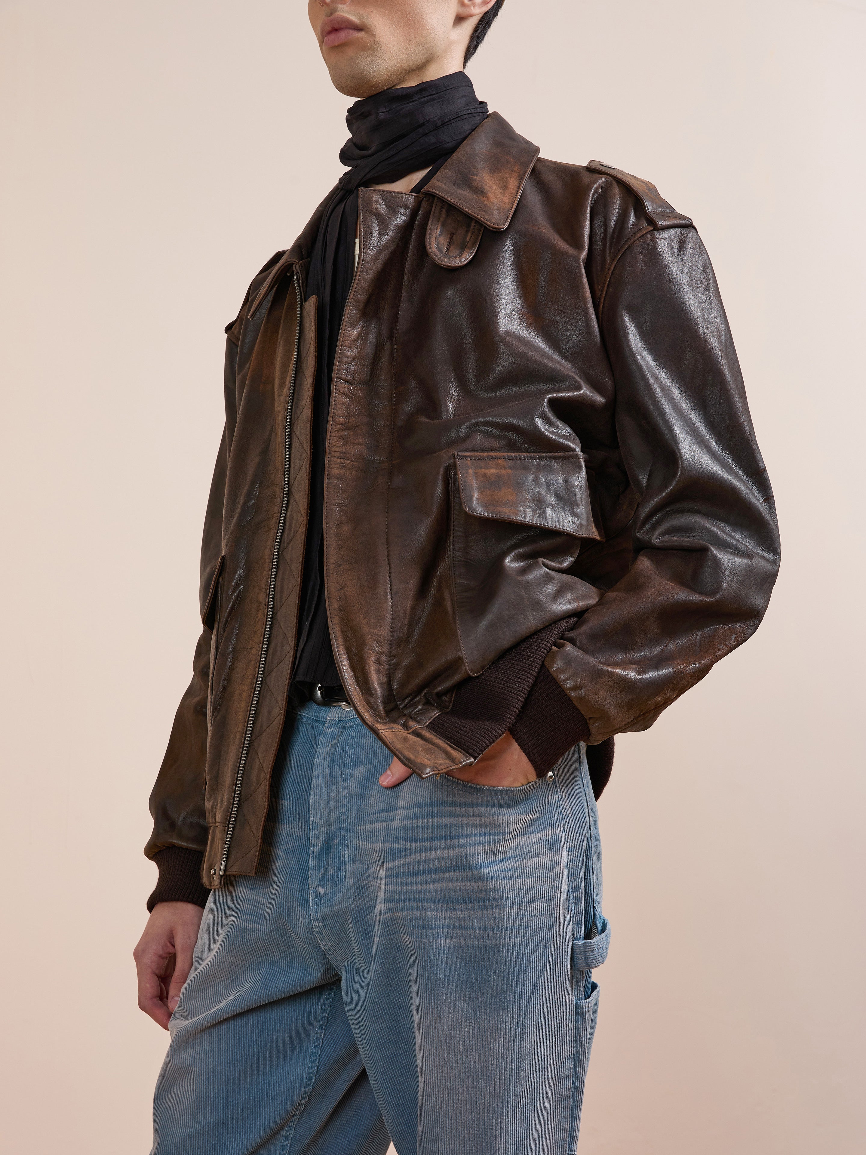 A person wears the FOUND Distressed Leather Flight Jacket, paired with a black scarf and light blue jeans, standing against a neutral background.