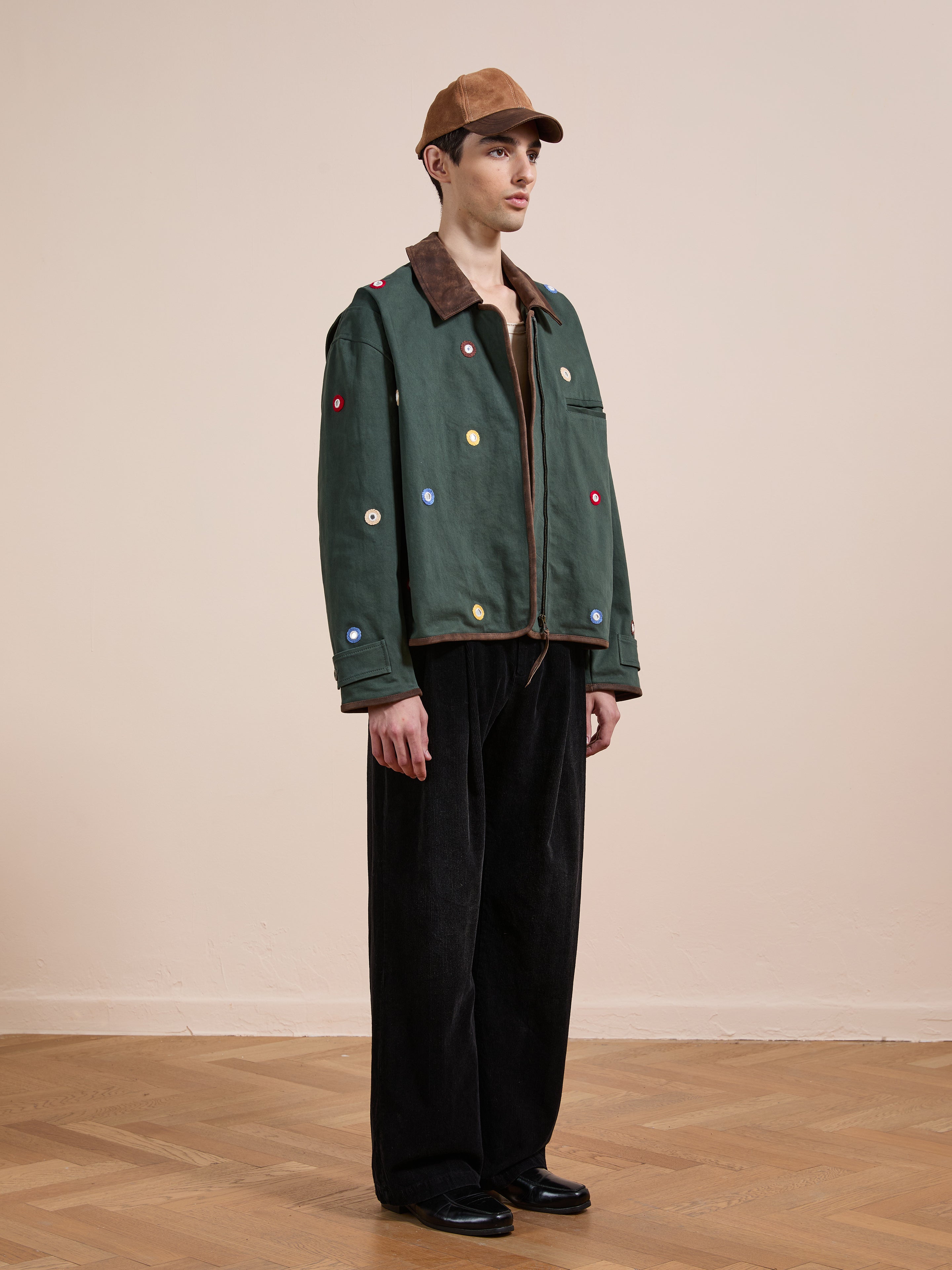 Indoors, a person wears Found’s Hand Embroidered Mirror Abhala Jacket with colorful patches, paired with a brown cap, brown shirt, loose black pants, and black shoes on a wooden floor.