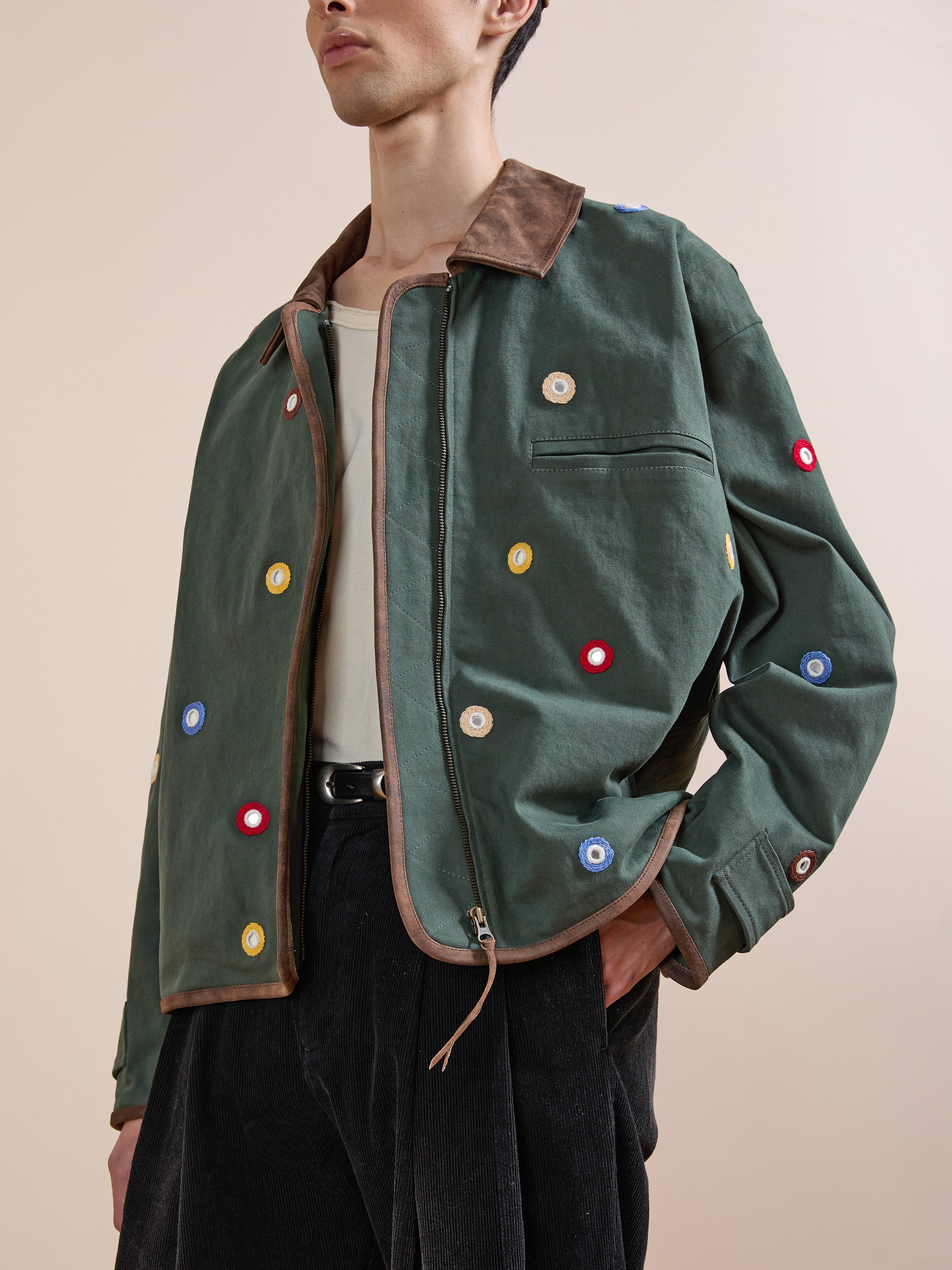 A person wears the Found Hand Embroidered Mirror Abhala Jacket—a handcrafted green cotton jacket with multicolored circular patches and a brown collar—over a beige shirt, paired with black pants.