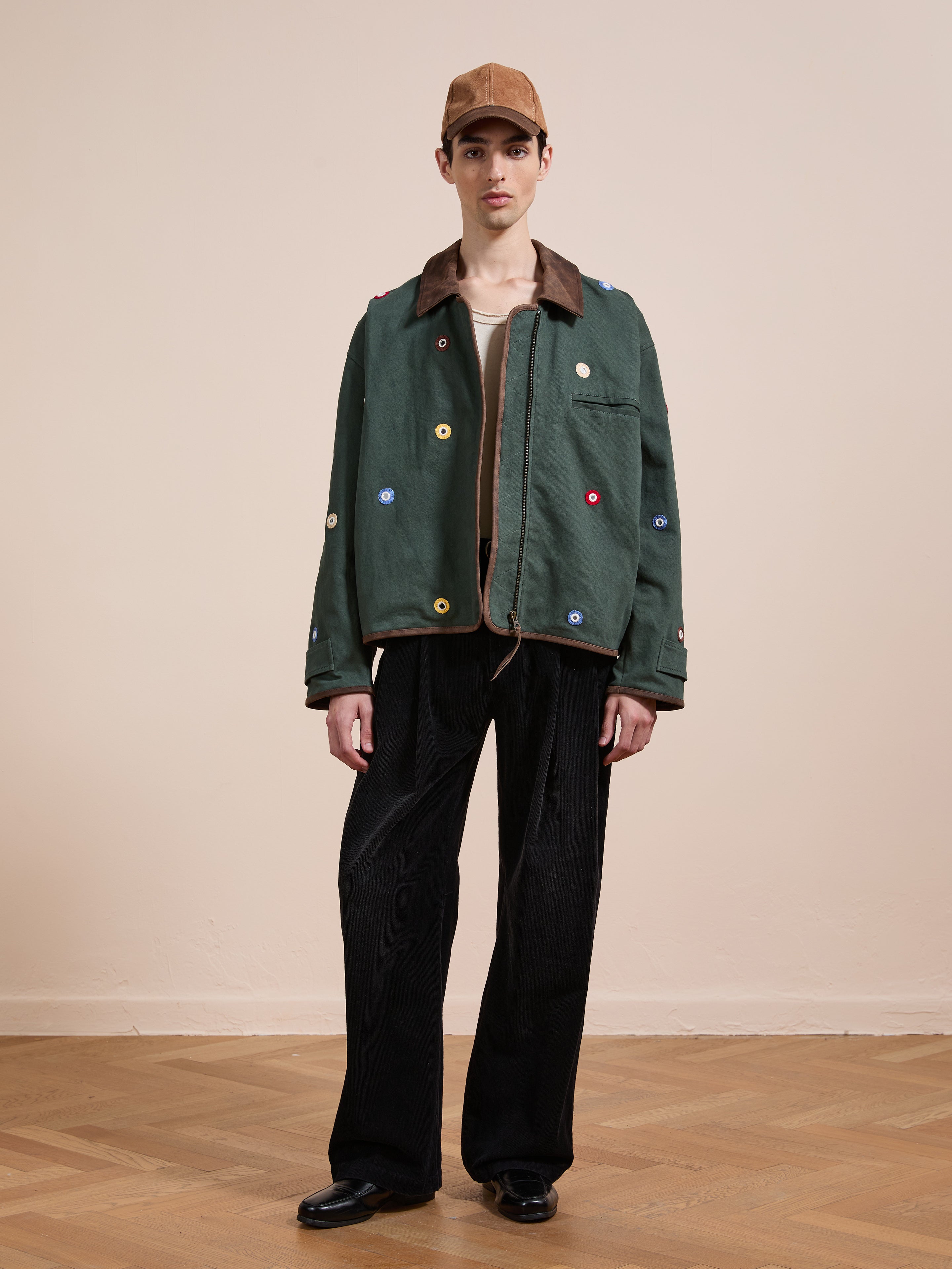 Indoors, a person wears Found's Hand Embroidered Mirror Abhala Jacket with colorful accents, paired with black wide-leg pants, a beige shirt, black shoes, and a brown cap.
