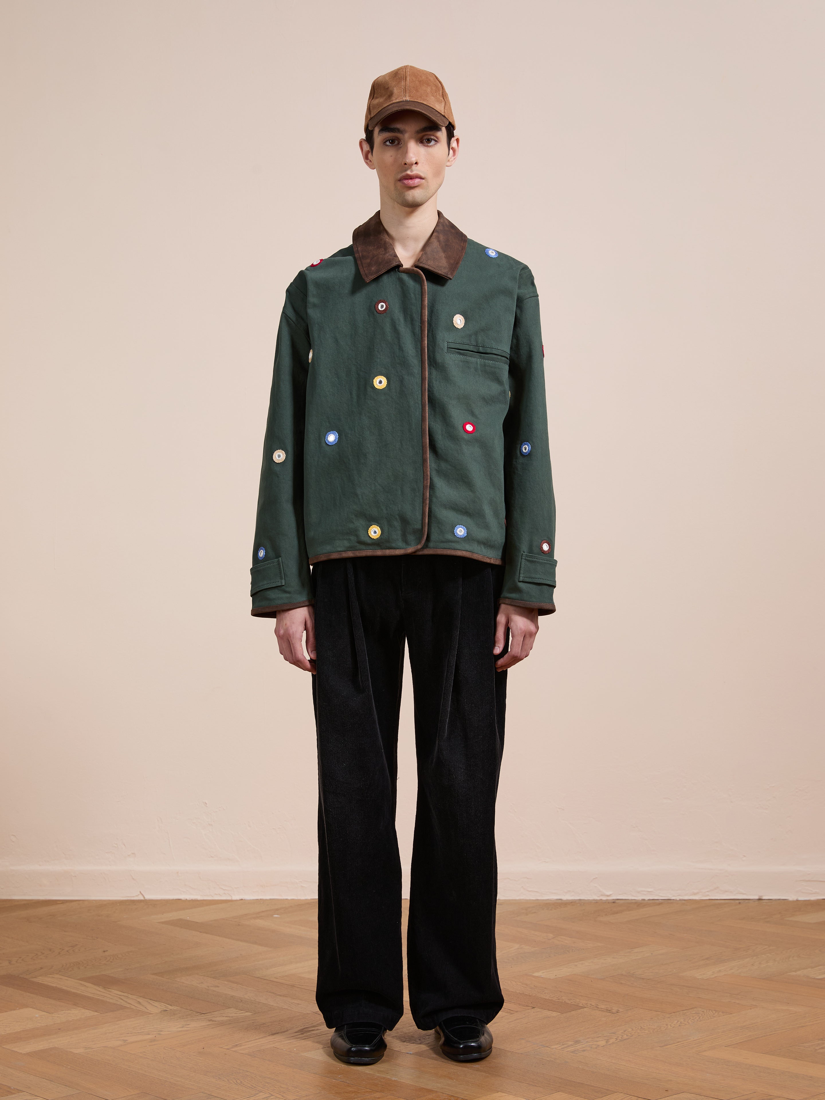 A person stands indoors in Found's Hand Embroidered Mirror Abhala Jacket—green with multicolored circular accents—paired with wide black pants, black shoes, and a brown cap against a plain background.