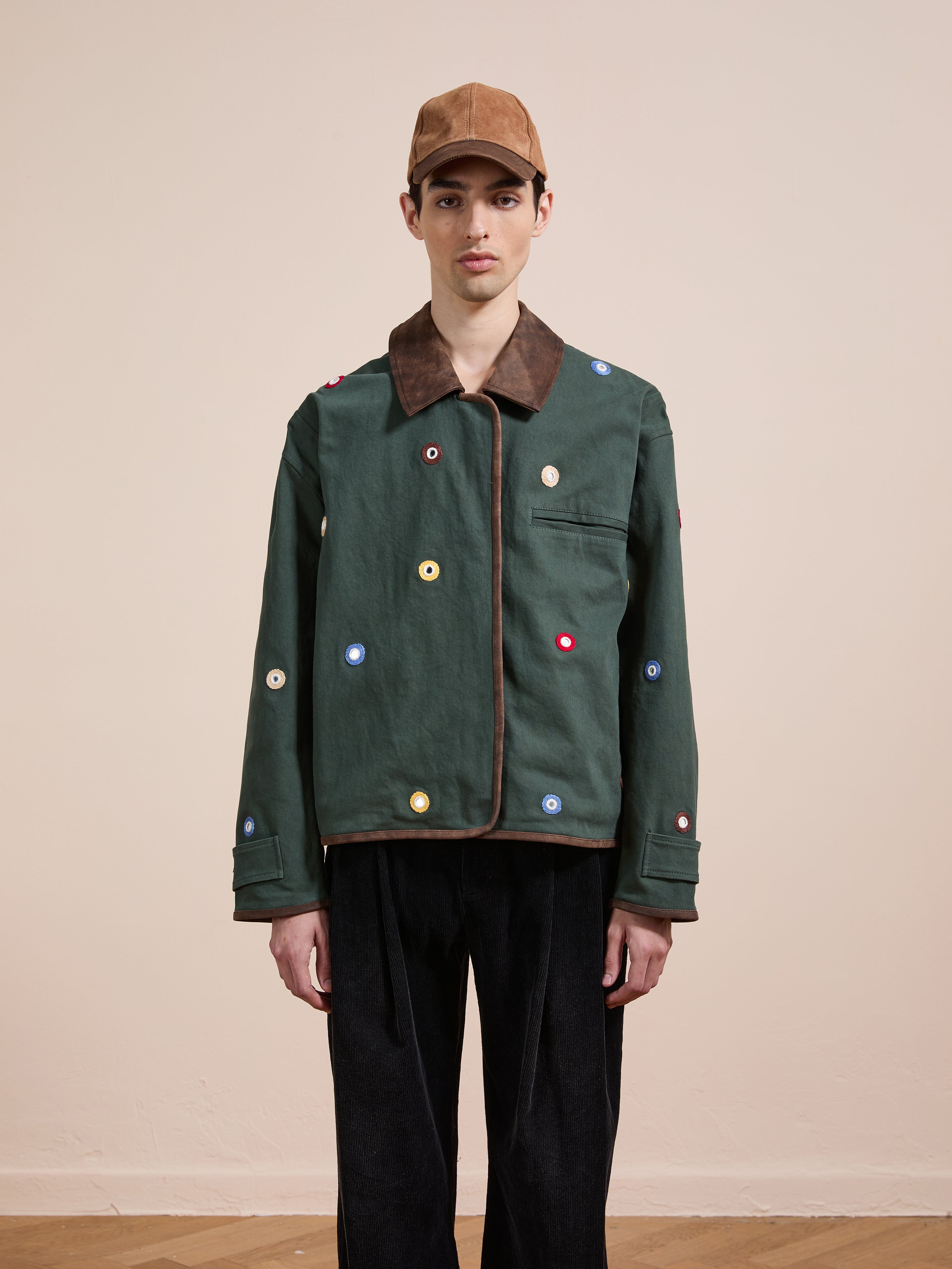 A person wearing a brown cap, black pants, and Found's Hand Embroidered Mirror Abhala Jacket stands against a plain beige wall.