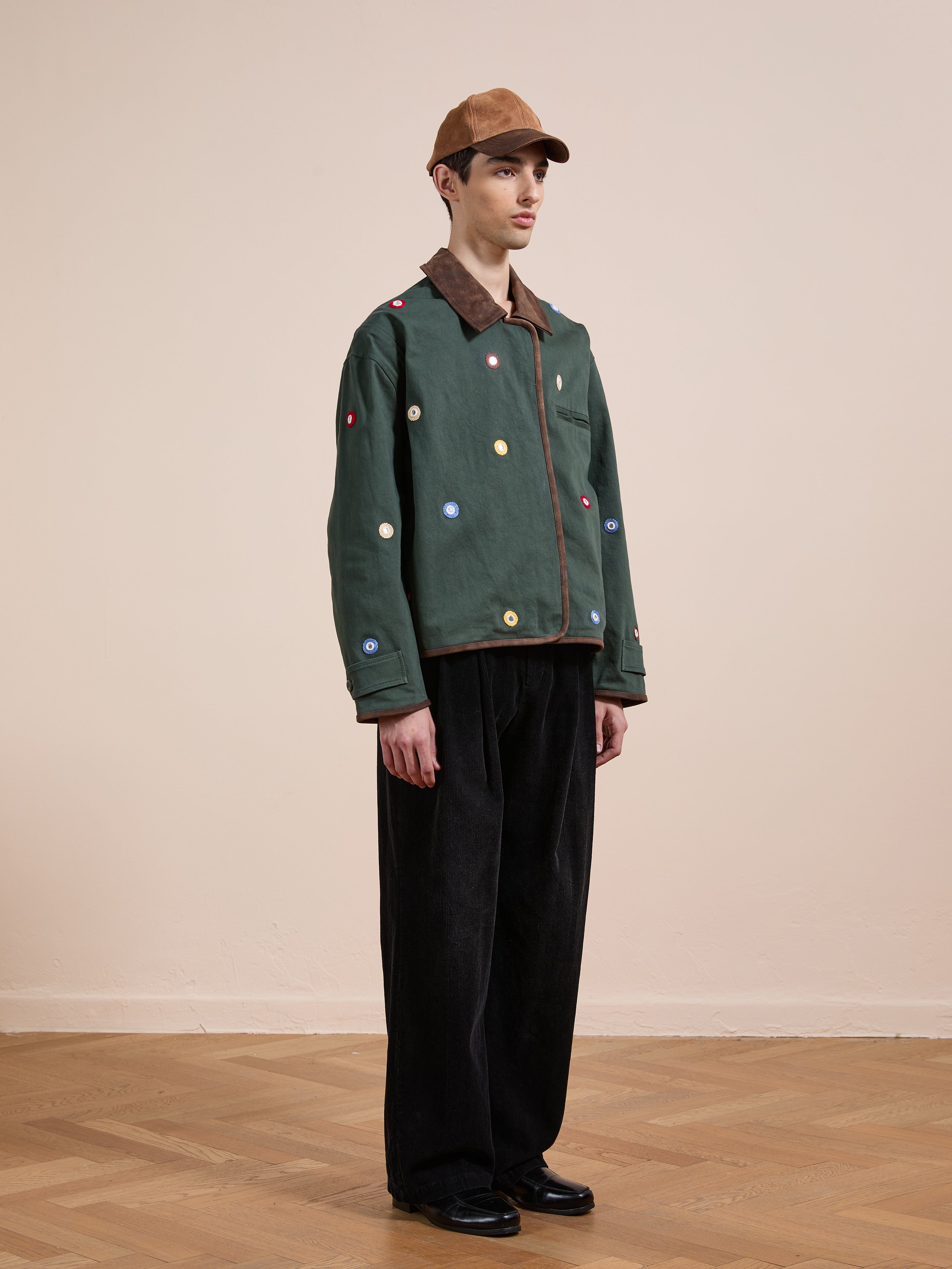 A person in a brown cap, black pants, and black shoes stands indoors on a wooden floor, showcasing the Found Hand Embroidered Mirror Abhala Jacket inspired by Pakistani Sheesha Kari with colorful circular patch details.