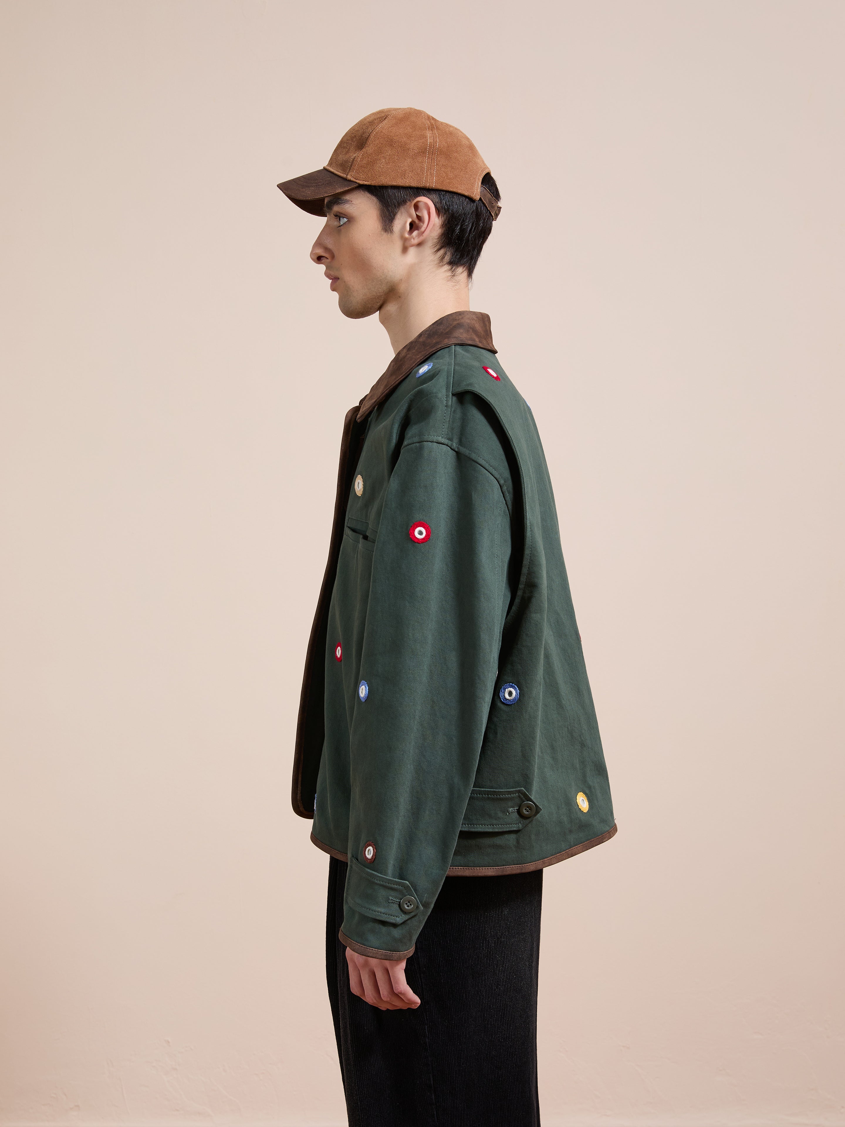 A man in profile wears Found's Hand Embroidered Mirror Abhala Jacket, featuring colorful circular patches, paired with a brown cap and dark pants, standing against a plain beige background.