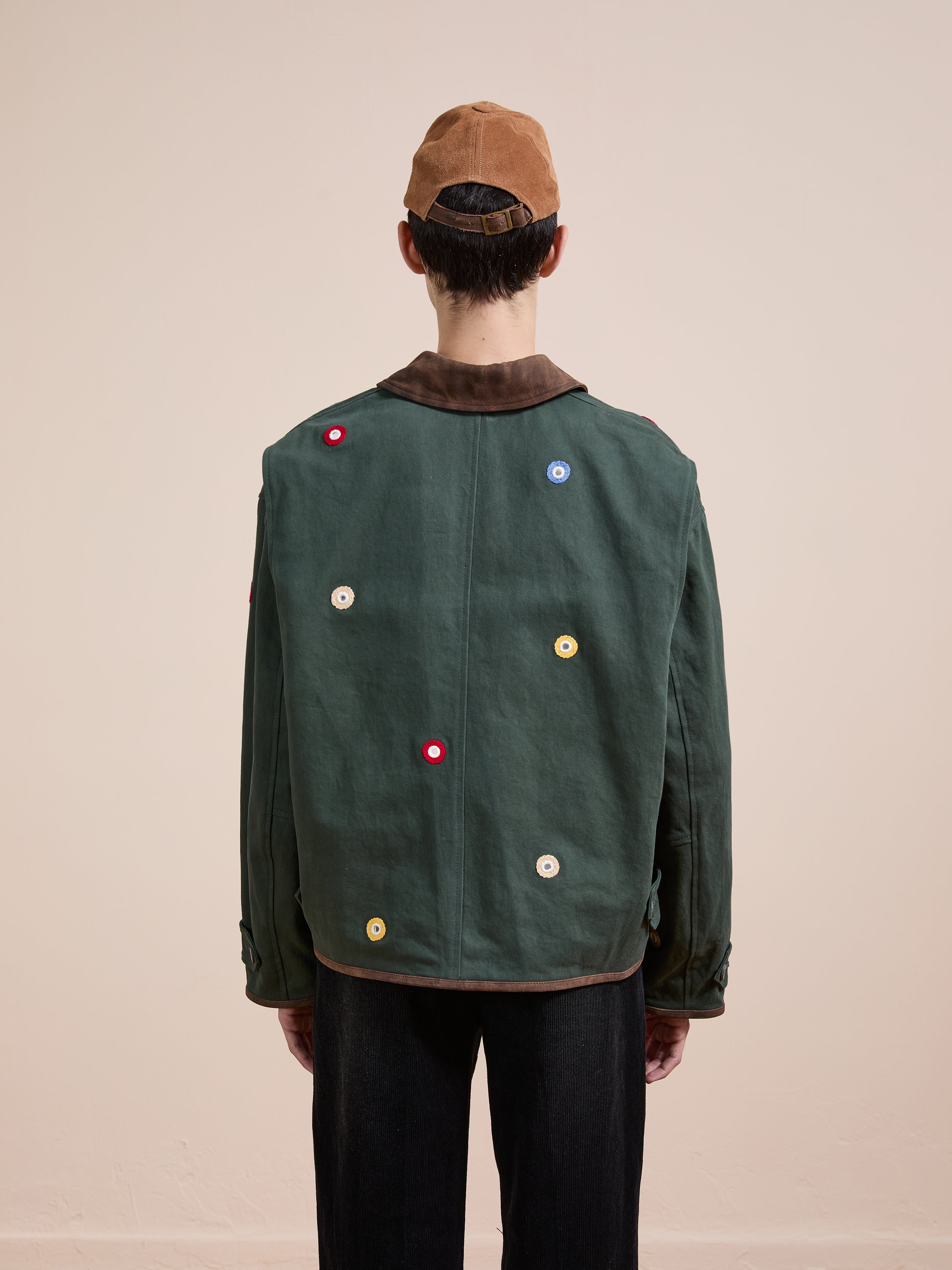 A person stands facing away, wearing Found's green Hand Embroidered Mirror Abhala Jacket with multicolored circular patches and a brown cap, set against a plain light background.
