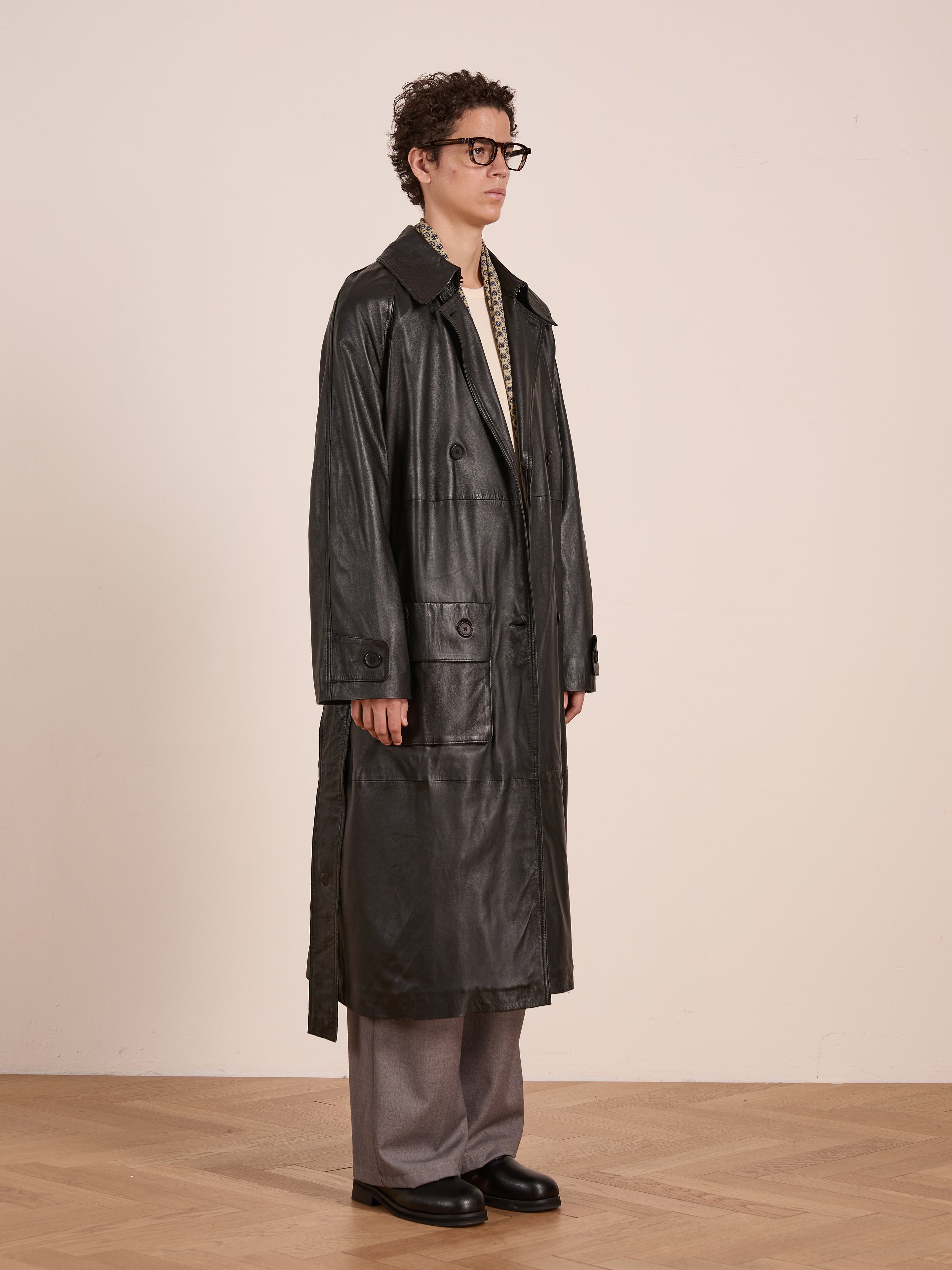A person in glasses wears the FOUND Leather Trench Coat, paired with grey wide-leg pants and black shoes, standing on a wooden floor against a plain, light-colored wall.