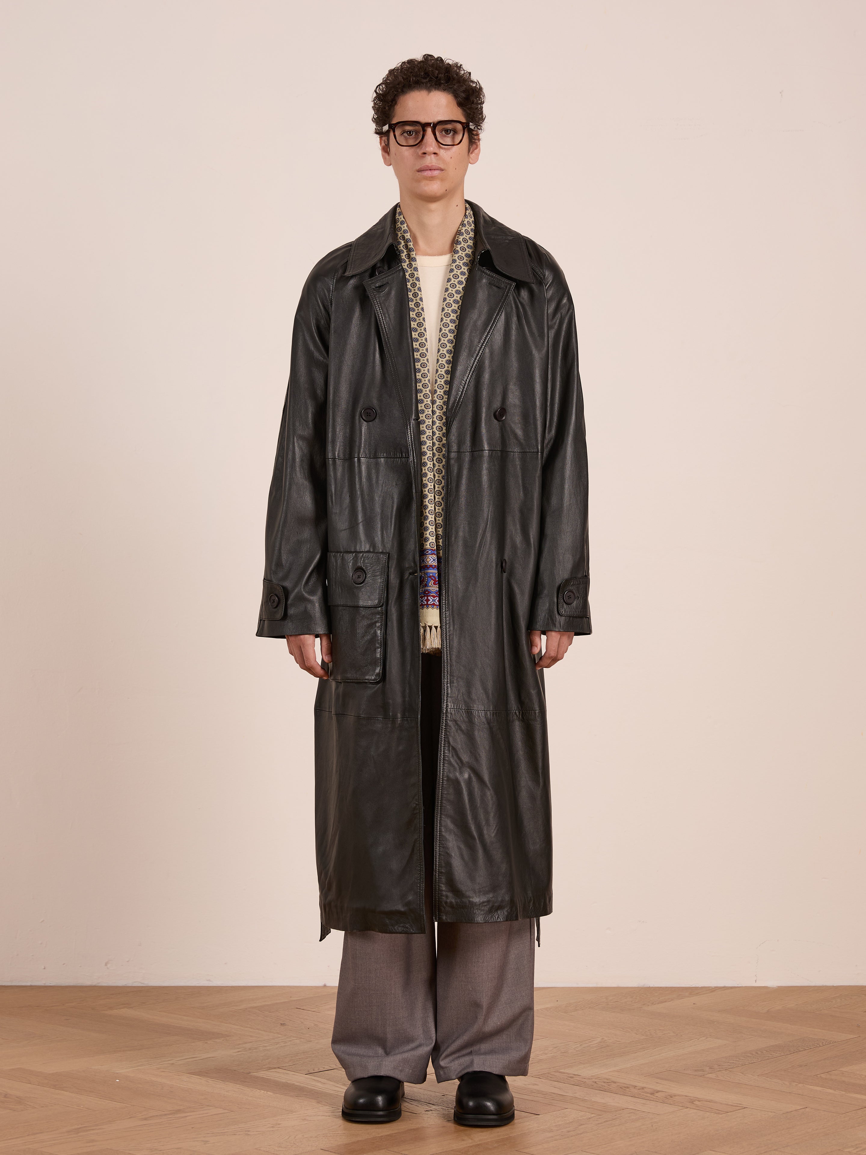 A person models the FOUND Leather Trench Coat over a light patterned sweater, wide gray pants, and black shoes while standing against a plain beige wall on a wooden floor.