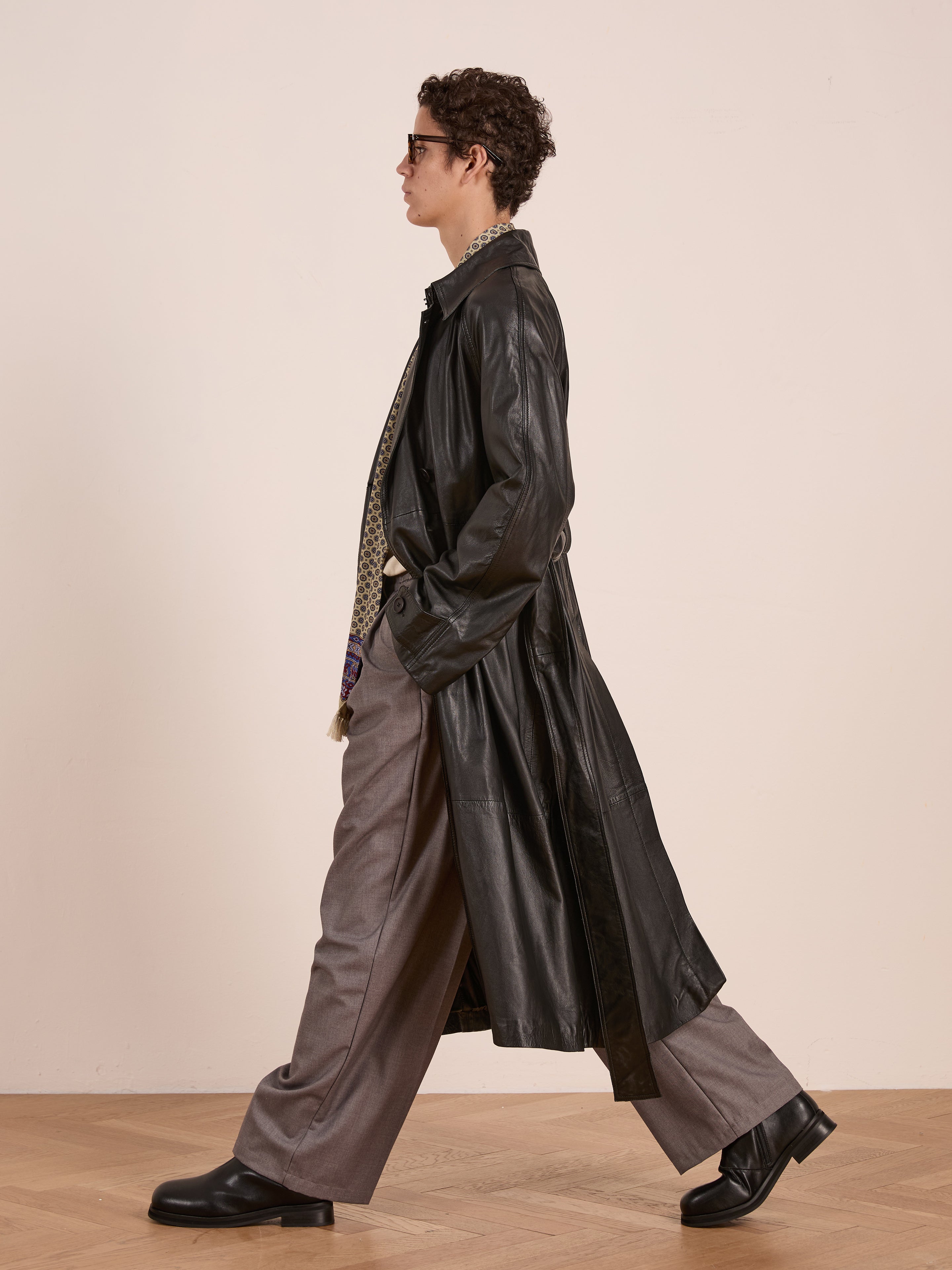 A person wearing the FOUND Leather Trench Coat, paired with gray trousers, black shoes, a patterned shirt, and sunglasses, walks indoors on a wooden floor.
