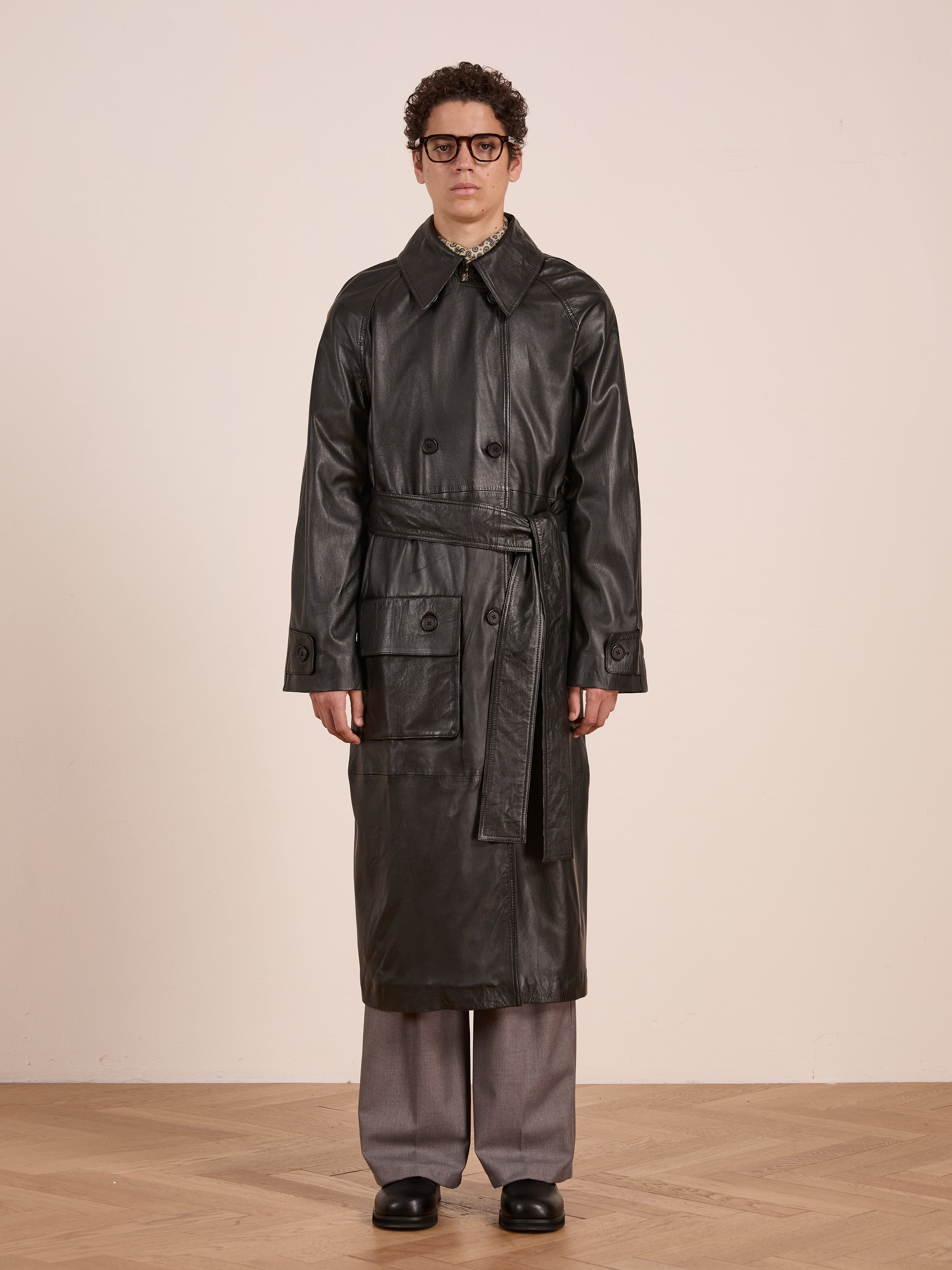 A person in glasses models the FOUND Leather Trench Coat, featuring a belt and large pockets, paired with gray trousers and black shoes, set against a plain light background.