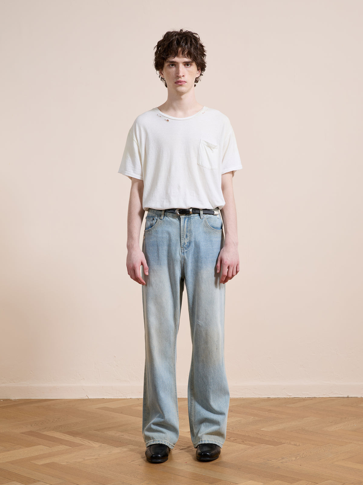 A person stands indoors against a plain wall, wearing a white t-shirt, Found Straight Leg Jeans, and black shoes on a wooden floor.
