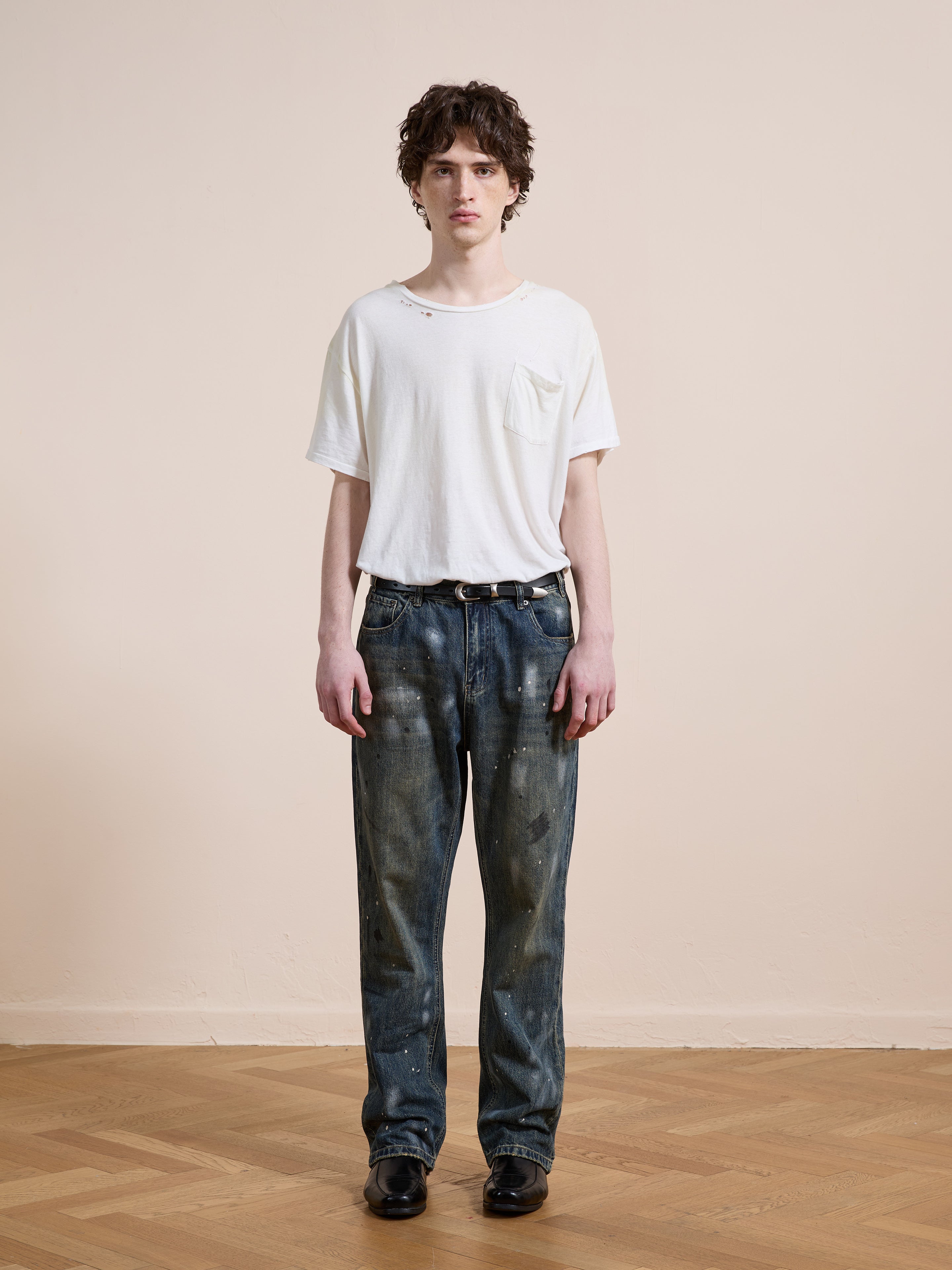 A young person faces forward in a distressed white t-shirt, FOUND Monet Paint Jeans in vintage blue wash (unisex relaxed fit), and black shoes, standing against a plain beige wall with a wooden floor.
