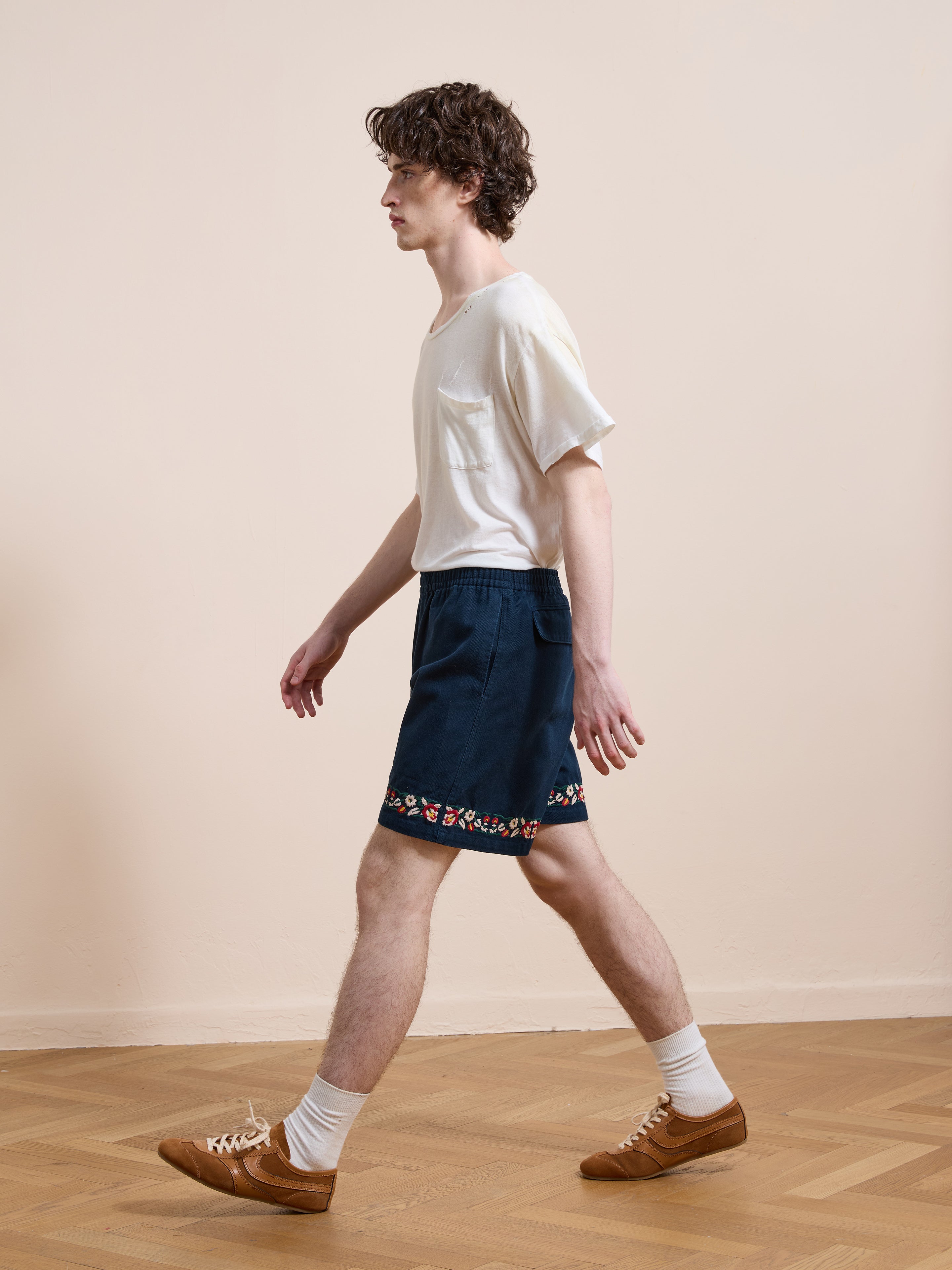 A person walks indoors wearing a white t-shirt, Found Horse Equine Twill Shorts with floral embroidery and a navy floral hem, white socks, and brown sneakers on a wooden floor against a plain beige wall.