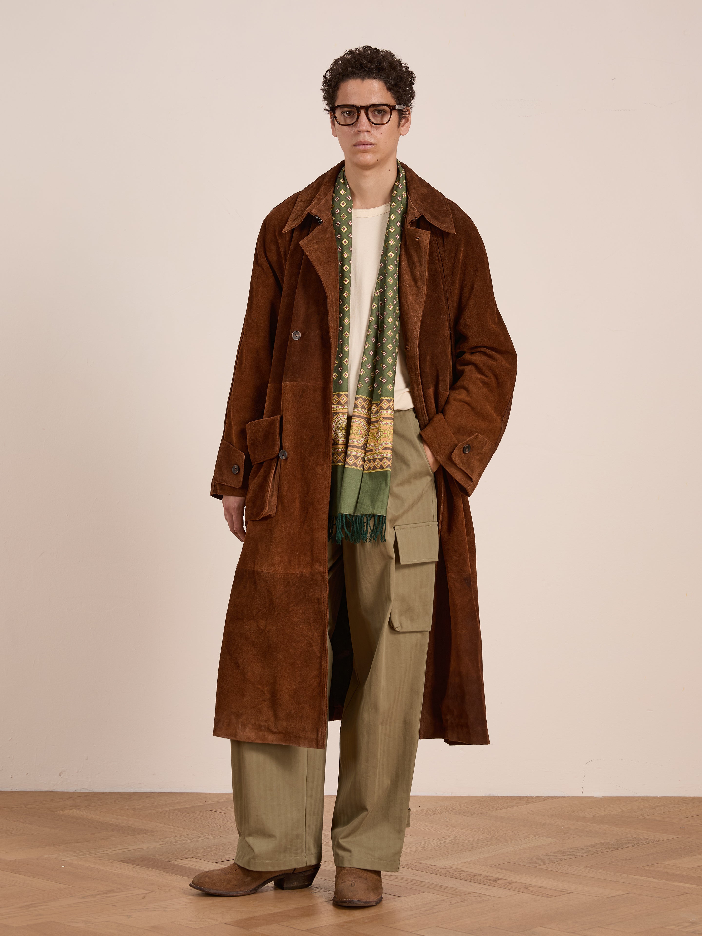 A person in FOUND’s Suede Leather Trench Coat, paired with a green patterned scarf, olive cargo pants, boots, and glasses, stands against a light wall. The mid-weight cow suede coat is ethically crafted in Pakistan.