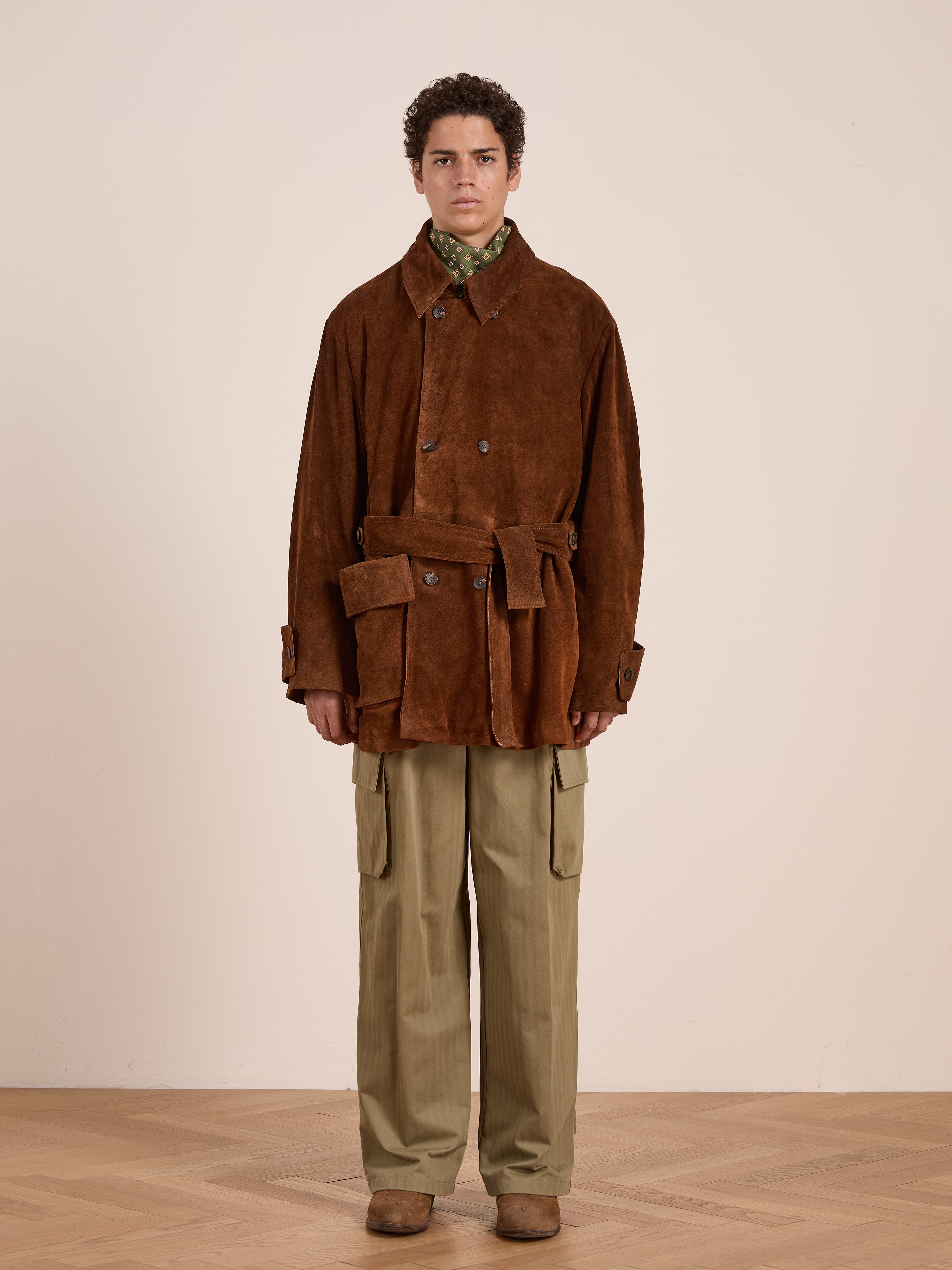 A person stands indoors against a light wall, wearing the FOUND Double Breasted Suede Leather Jacket—an ethically produced, unisex relaxed-fit brown suede coat—over a green shirt, beige cargo pants, and brown shoes.