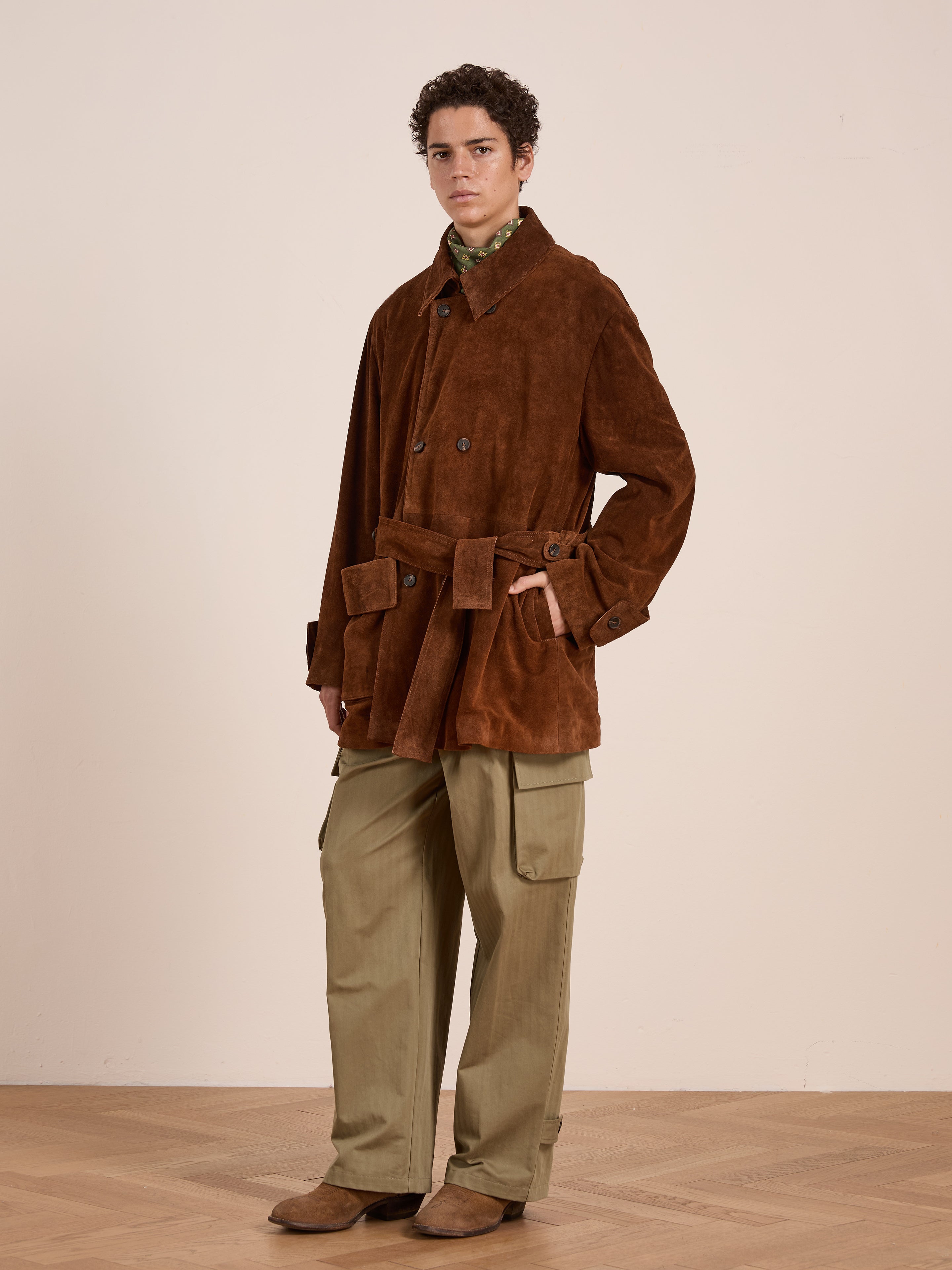 A person stands indoors against a plain light wall, wearing FOUND’s Double Breasted Suede Leather Jacket with front pockets and a belt, paired with green cargo pants and brown shoes.