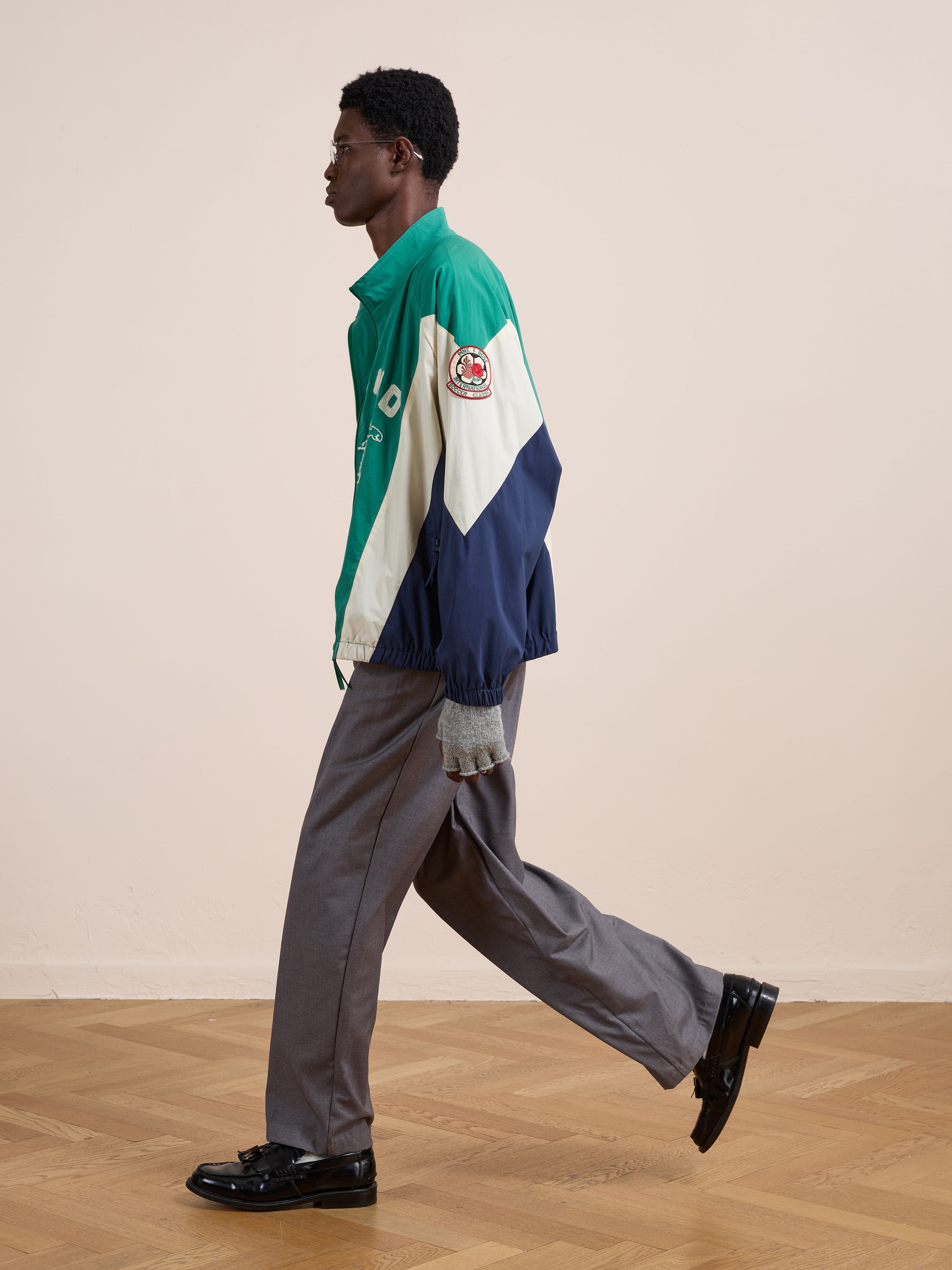 A person wearing a green, cream, and navy FOUND Crestline Sport Zip Jacket with a patch, gray pants, and black shoes walks indoors on a parquet floor, exuding vintage sportswear style.