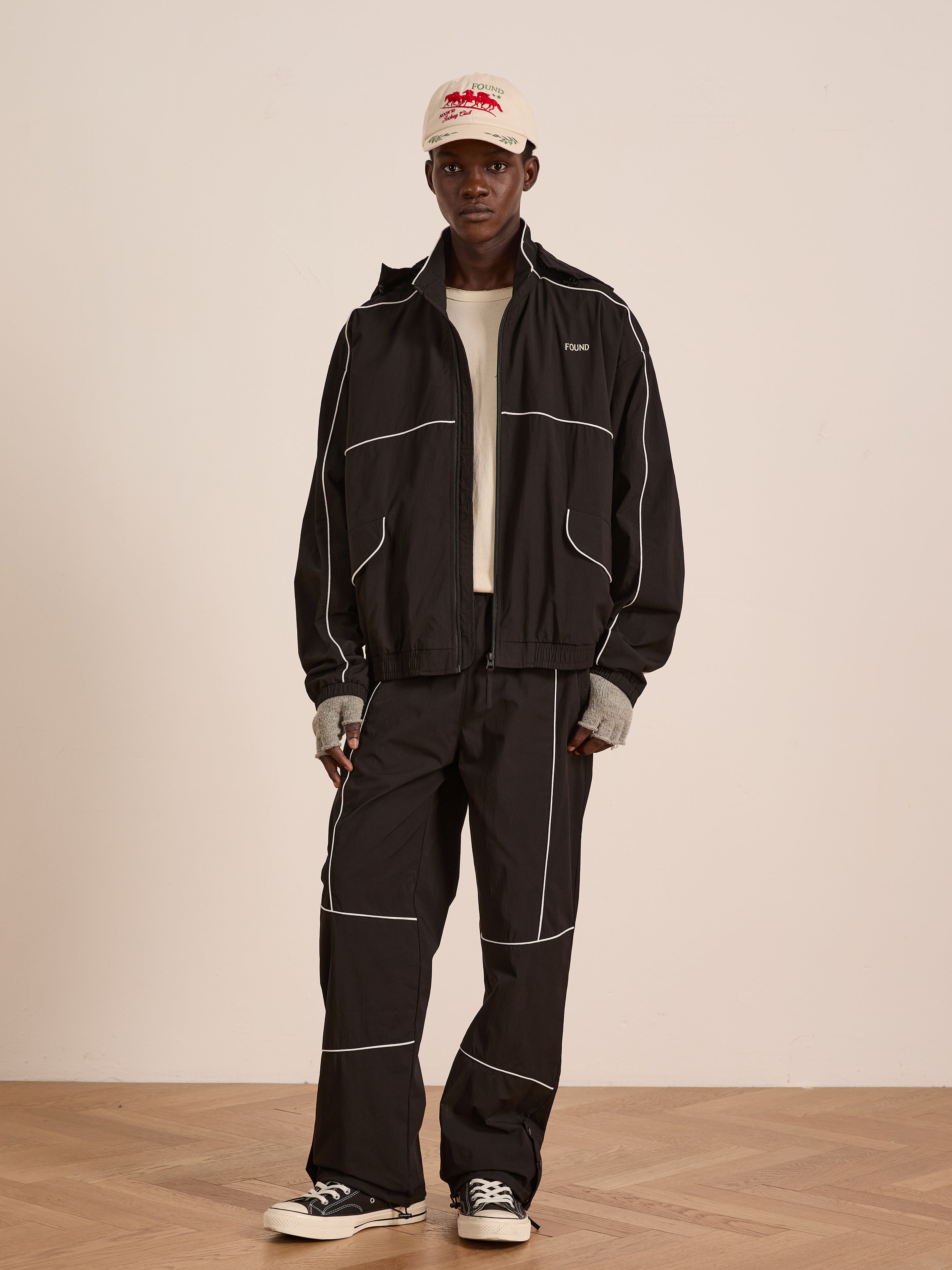 Indoors, a person wears the FOUND Arena Stripe Track Jacket in black with white piping, paired with a beige T-shirt, cap, fingerless gloves, and black lightweight technical sneakers.