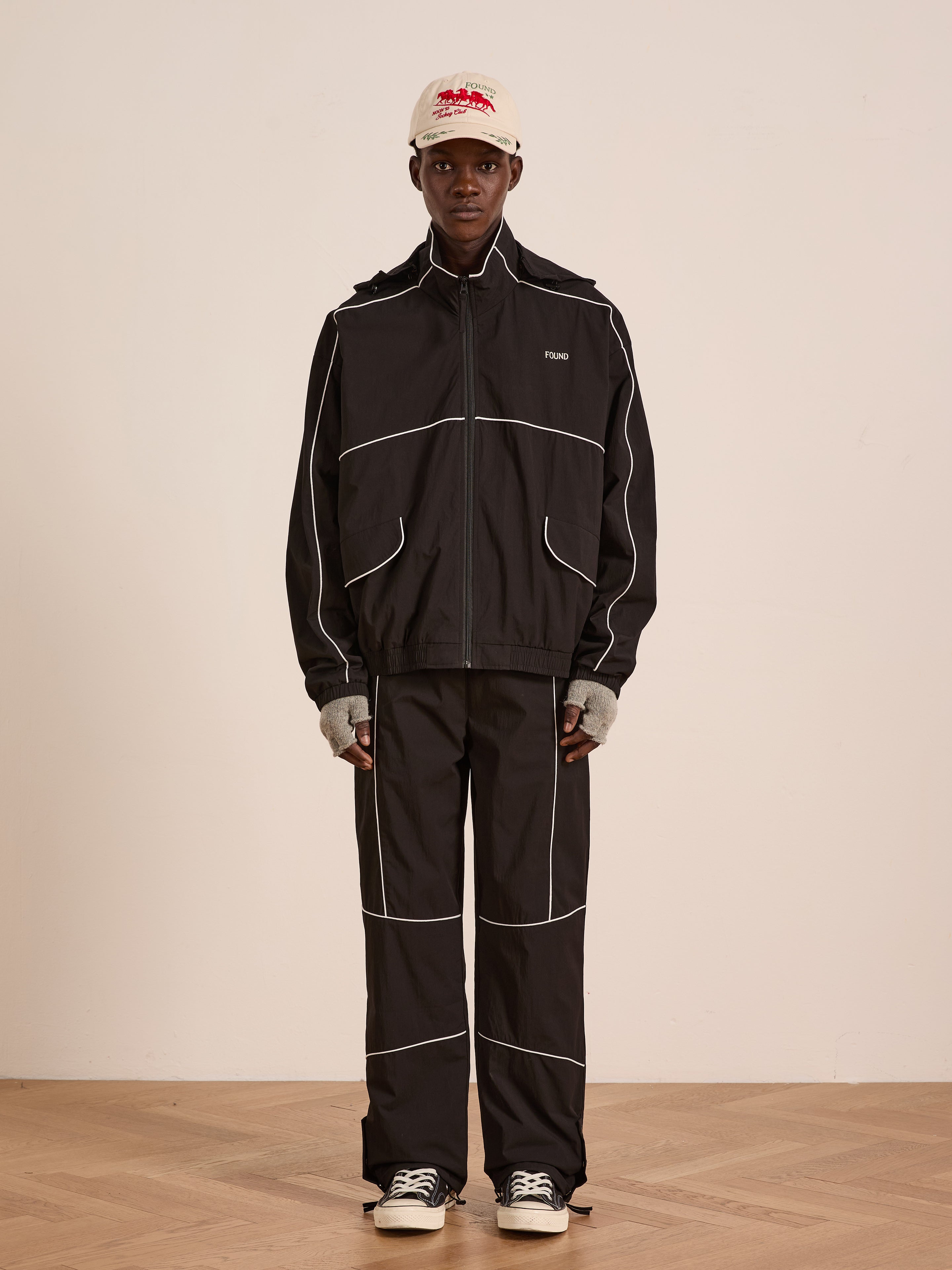 A person stands indoors in Found’s Arena Stripe Track Pants—a black pair with white piping and an adjustable waistband—styled with a beige cap, gray fingerless gloves, and black-and-white sneakers against a plain background.