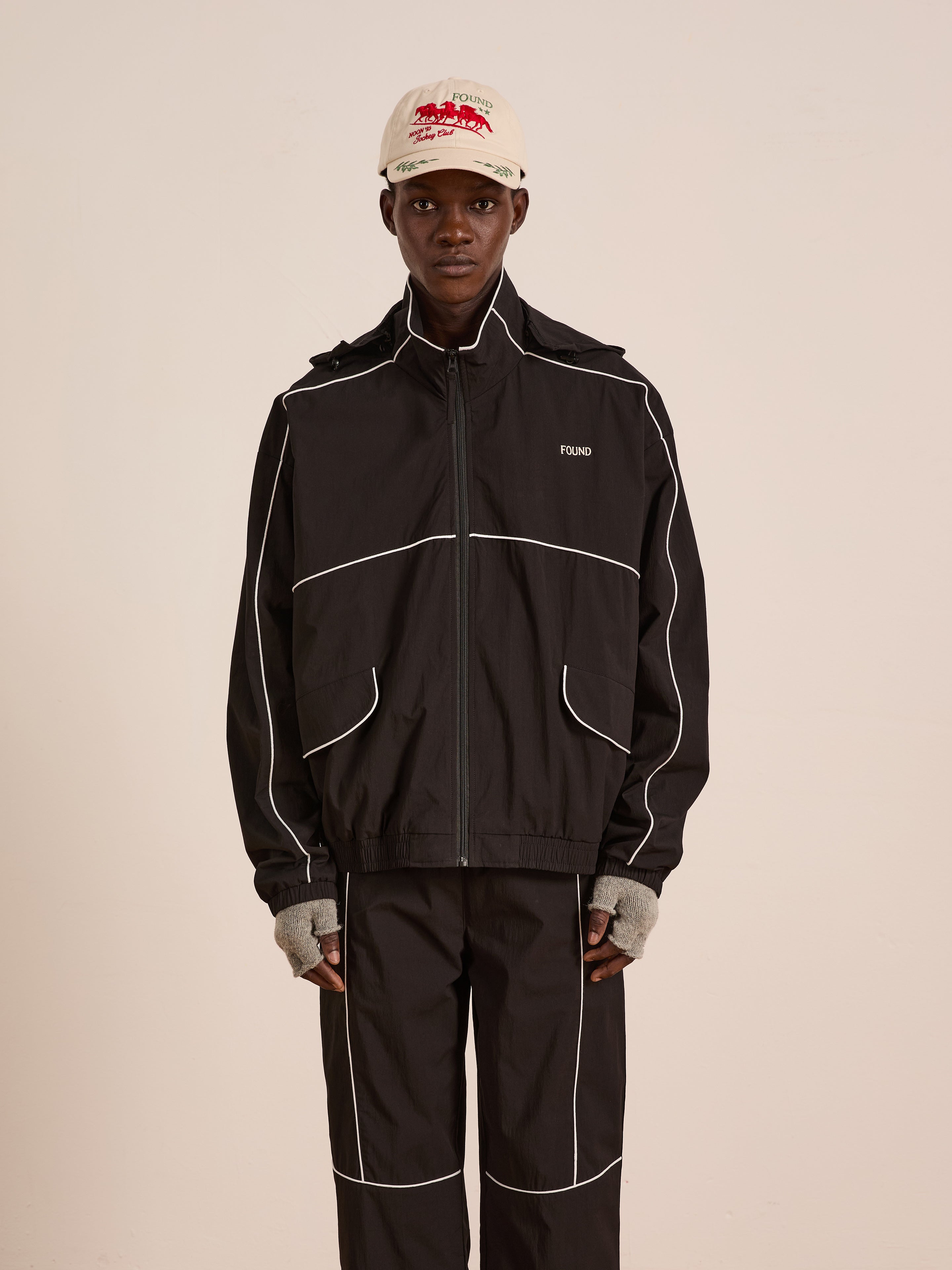 A person wears the FOUND Arena Stripe Track Jacket in black with white piping, matching pants, a white cap with red text, and gray fingerless gloves, standing against a plain light background. The jacket is made from lightweight technical fabric.