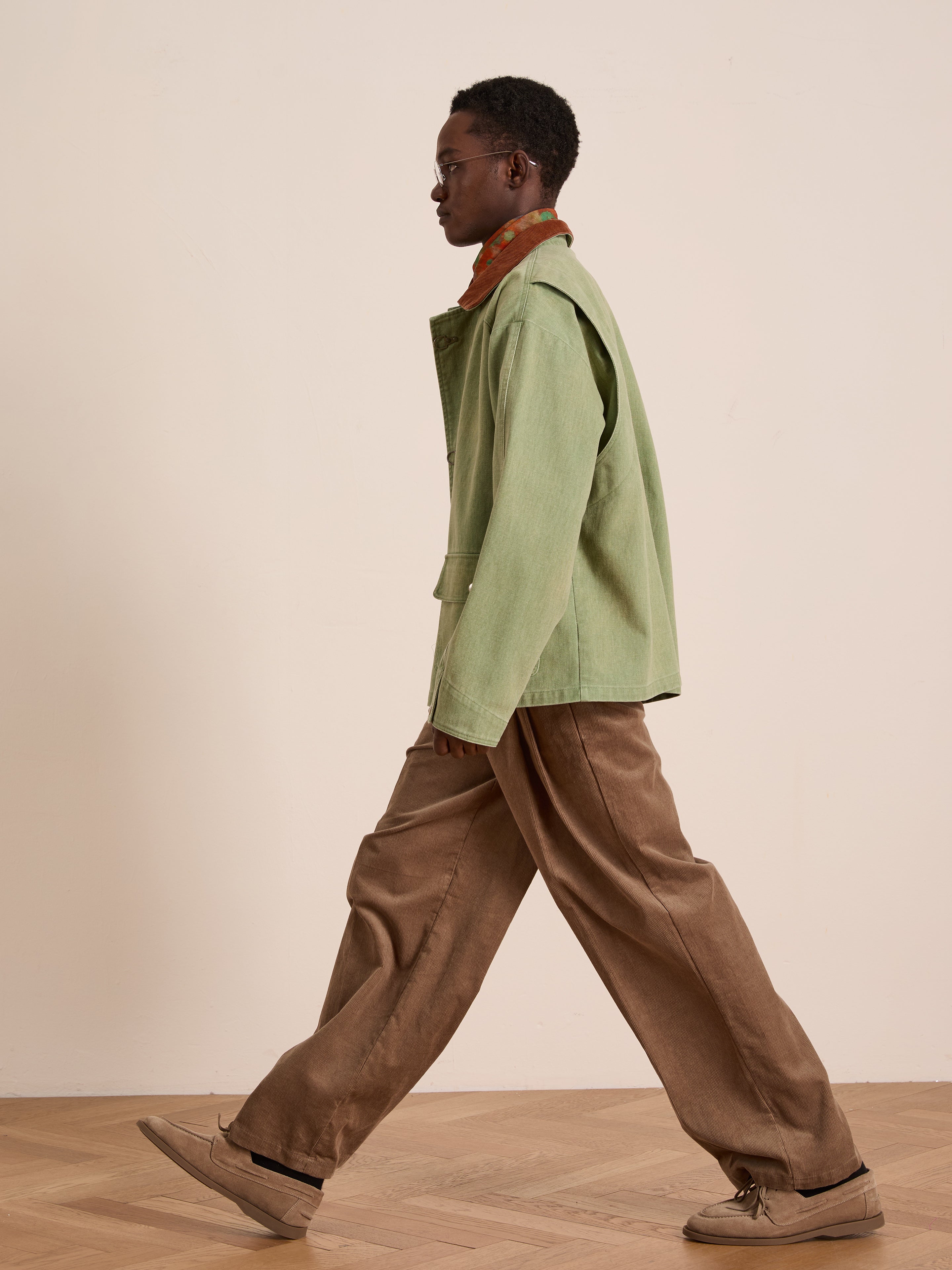 A person wears the Found Washed Fireman Clasp Workshop Jacket with a corduroy collar, brown pants, and tan shoes while walking indoors on a wooden floor against a plain beige wall.
