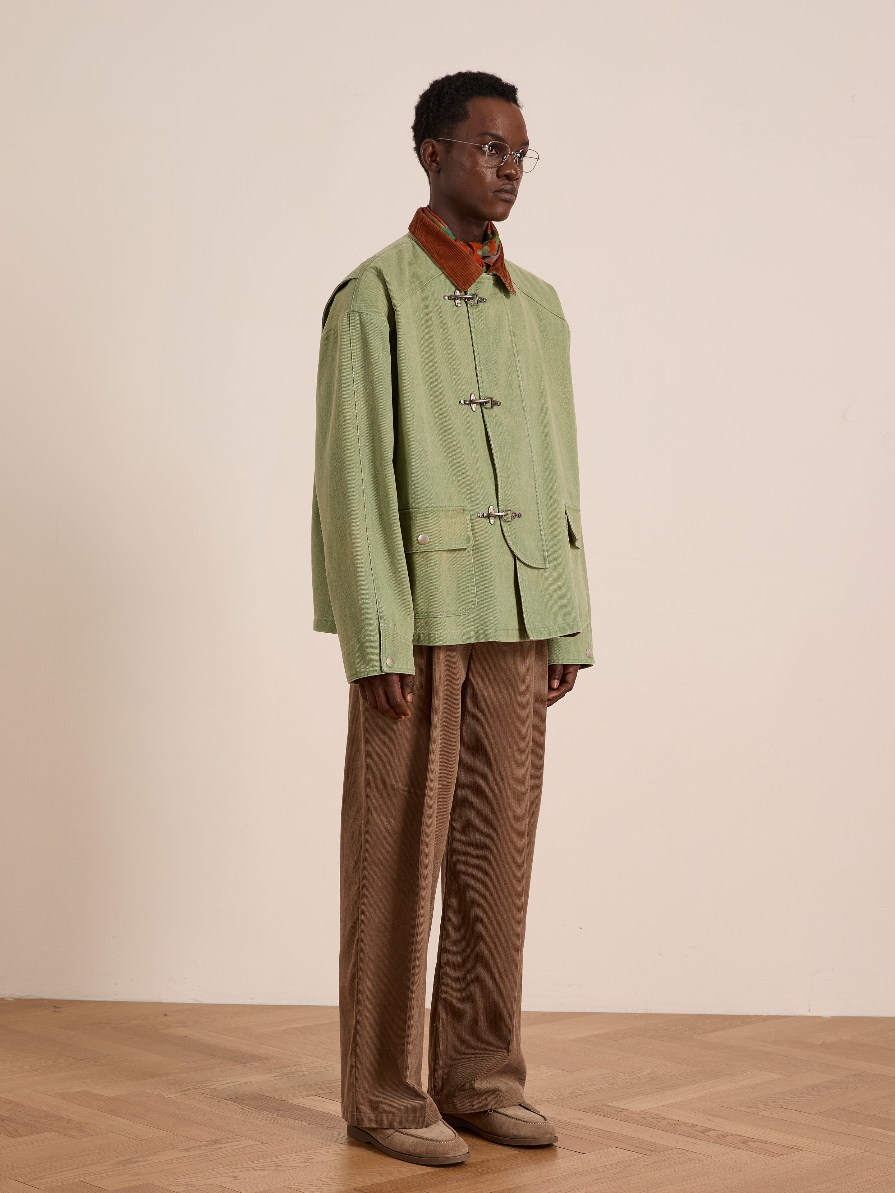 A person stands indoors against a light background wearing the Found Washed Fireman Clasp Workshop Jacket in light green with a brown corduroy collar, loose brown pants, brown shoes, and round glasses.