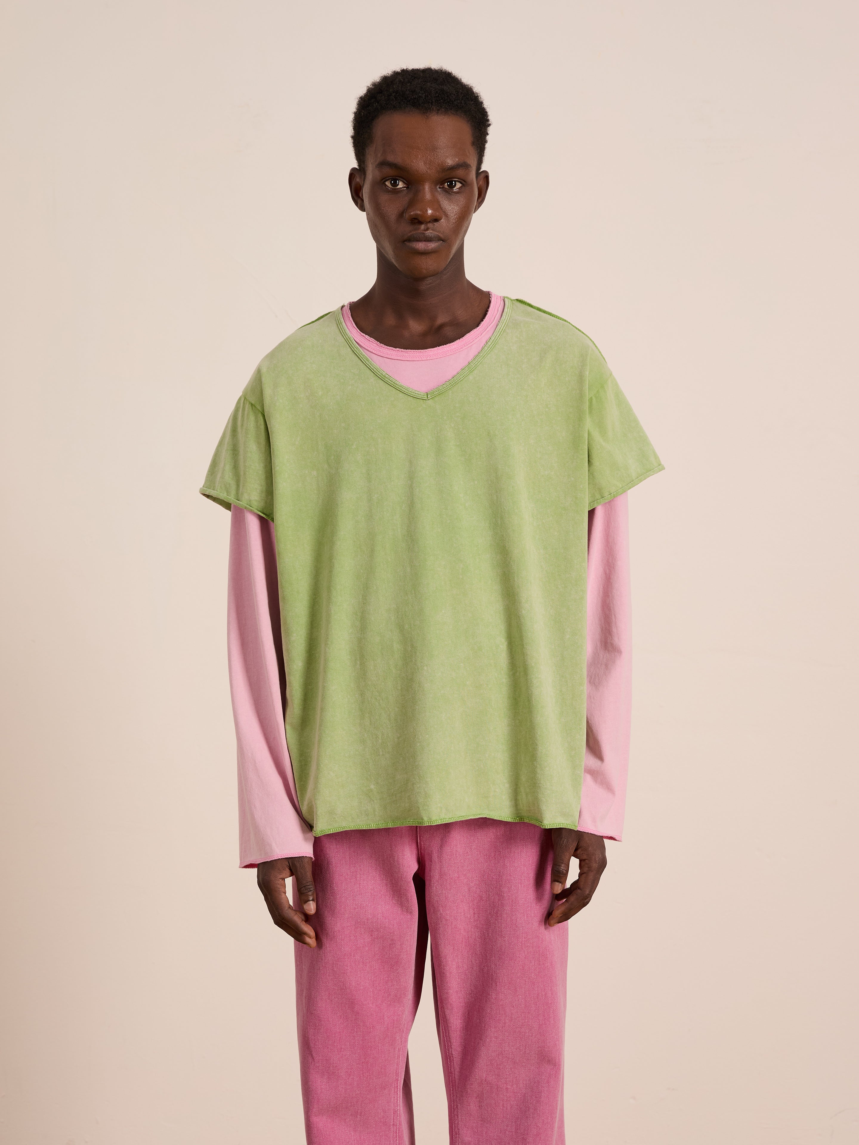 A person stands against a plain background wearing the FOUND Double Layer LS Tee—a green short-sleeve top layered over a pink unisex long sleeve tee—paired with pink pants.