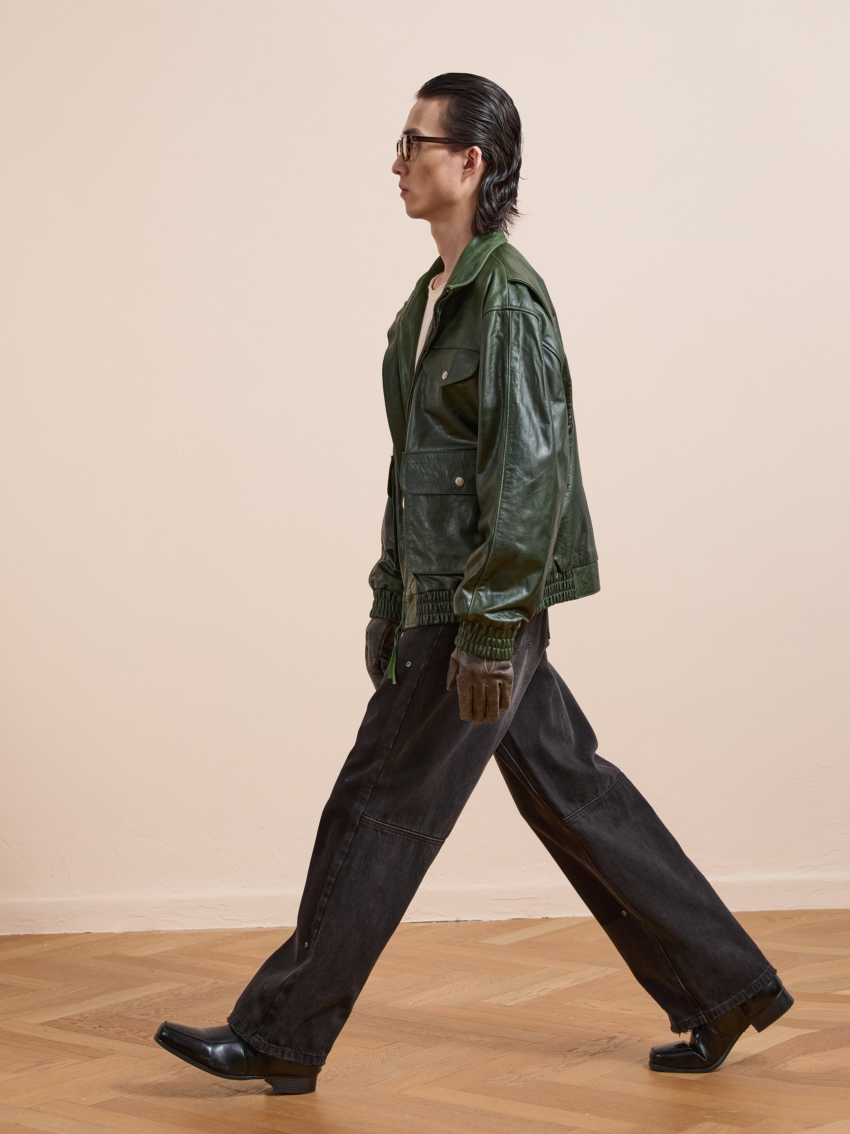 A person in sunglasses, the FOUND Forest Leather Bomber Jacket, dark wide-leg pants, and black shoes walks indoors on a wooden floor against a plain beige wall.
