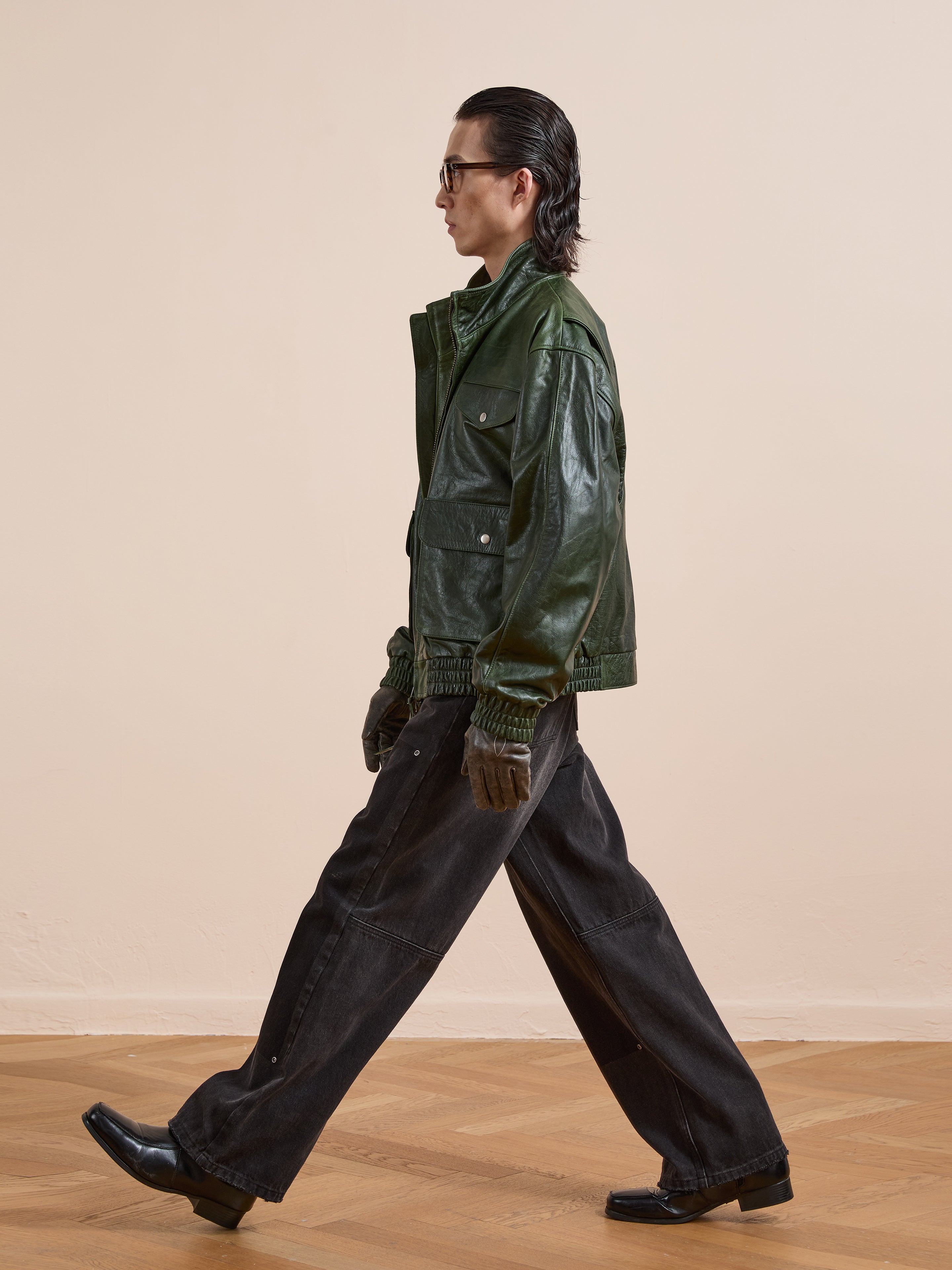 A person wearing the FOUND Forest Leather Bomber Jacket, wide-leg black pants, gloves, and black shoes walks across a wooden floor with a plain beige background.
