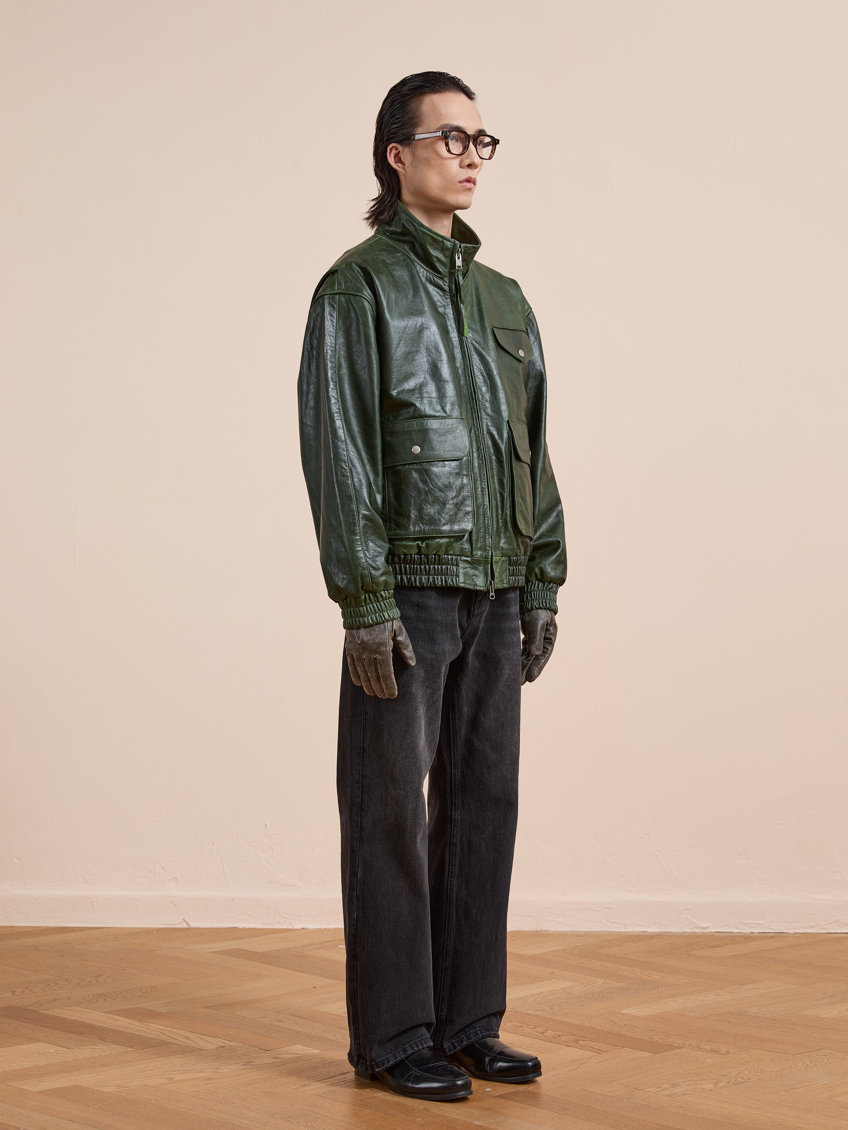 A person stands indoors against a beige wall and wooden floor, wearing the FOUND Forest Leather Bomber Jacket, dark gray pants, gloves, black shoes, and black glasses.