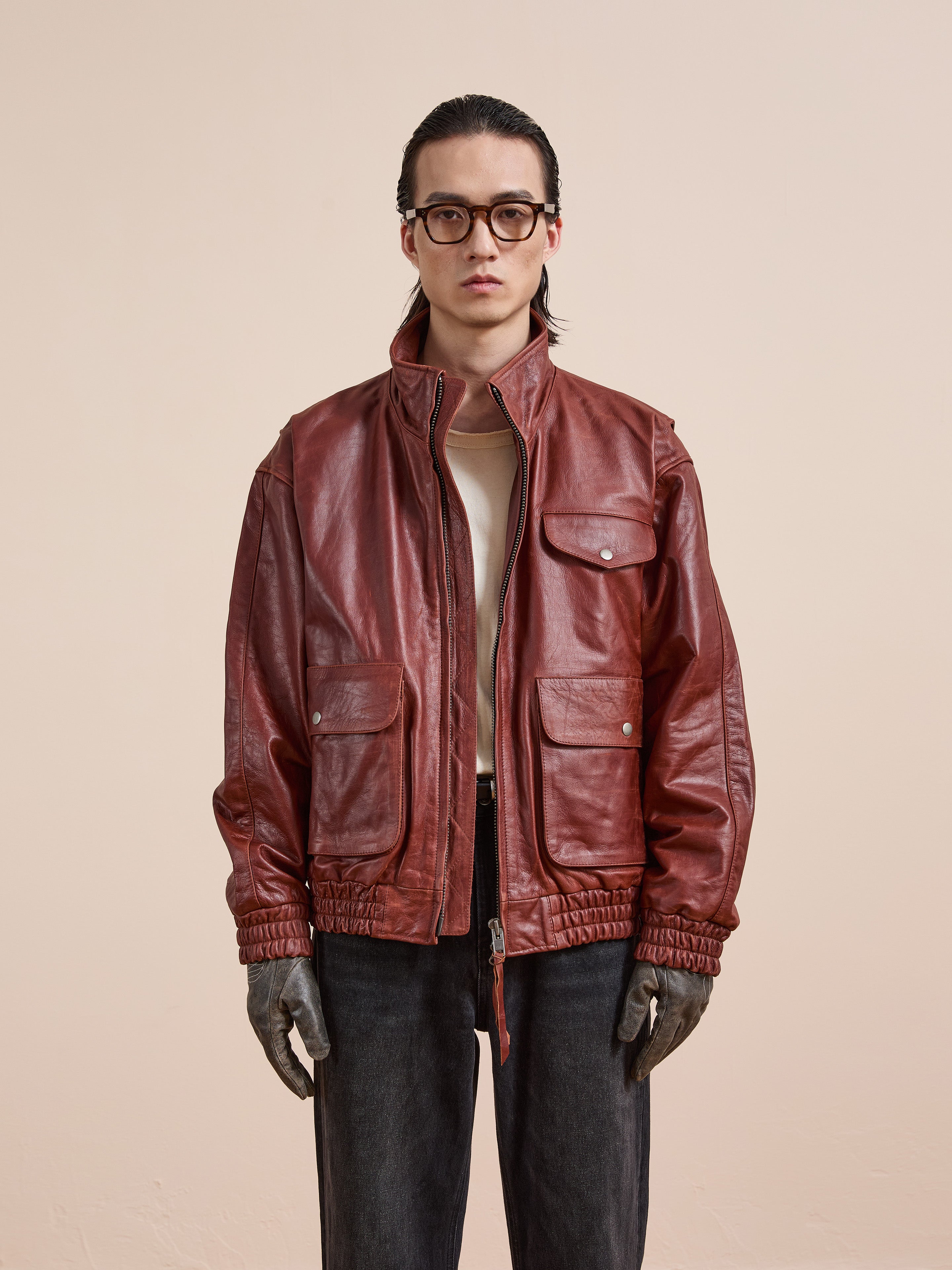 A person wearing glasses, a tan shirt, the Found Redwood Leather Bomber Jacket with multiple pockets, dark jeans, and gloves stands against a plain light background.