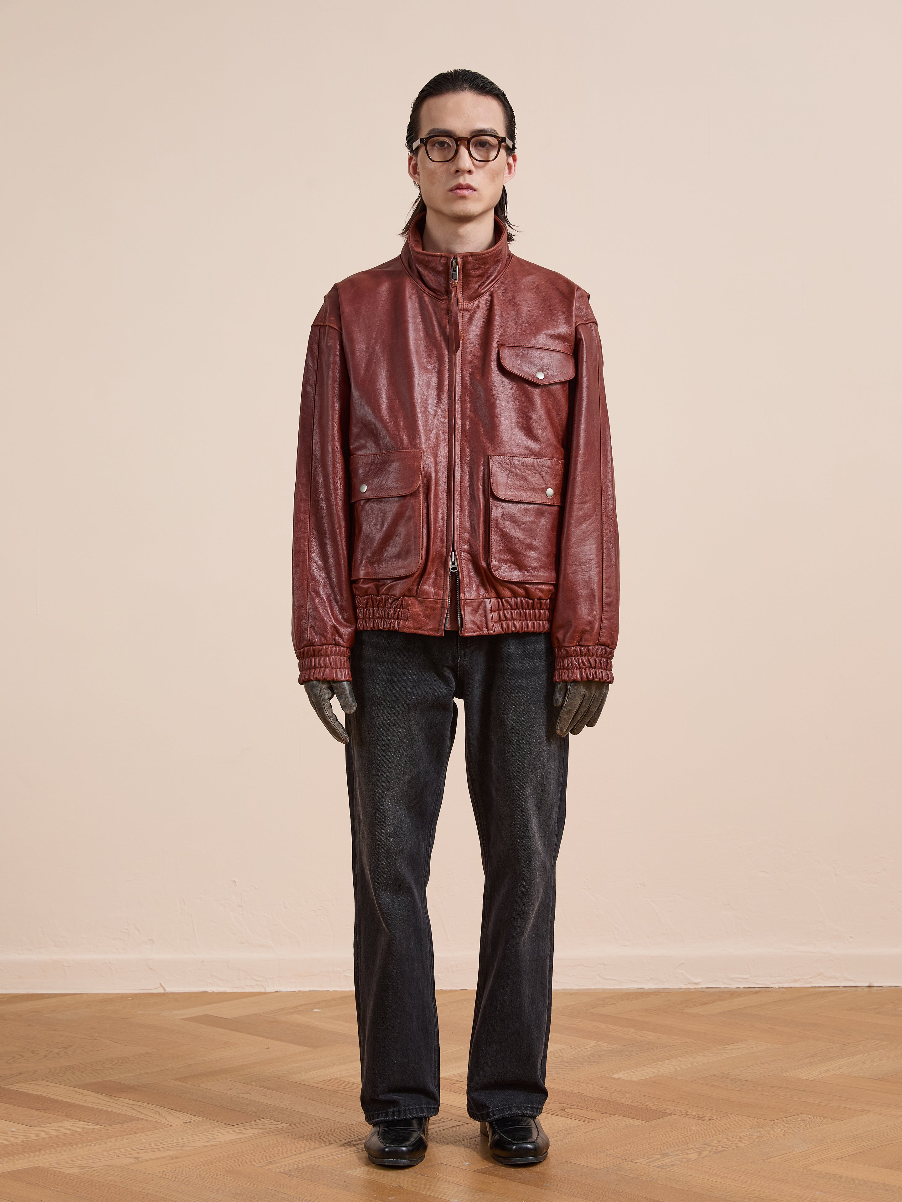 A person stands indoors facing forward against a beige wall, wearing Found's Redwood Leather Bomber Jacket in red, paired with dark jeans, black shoes, gray gloves, and glasses.