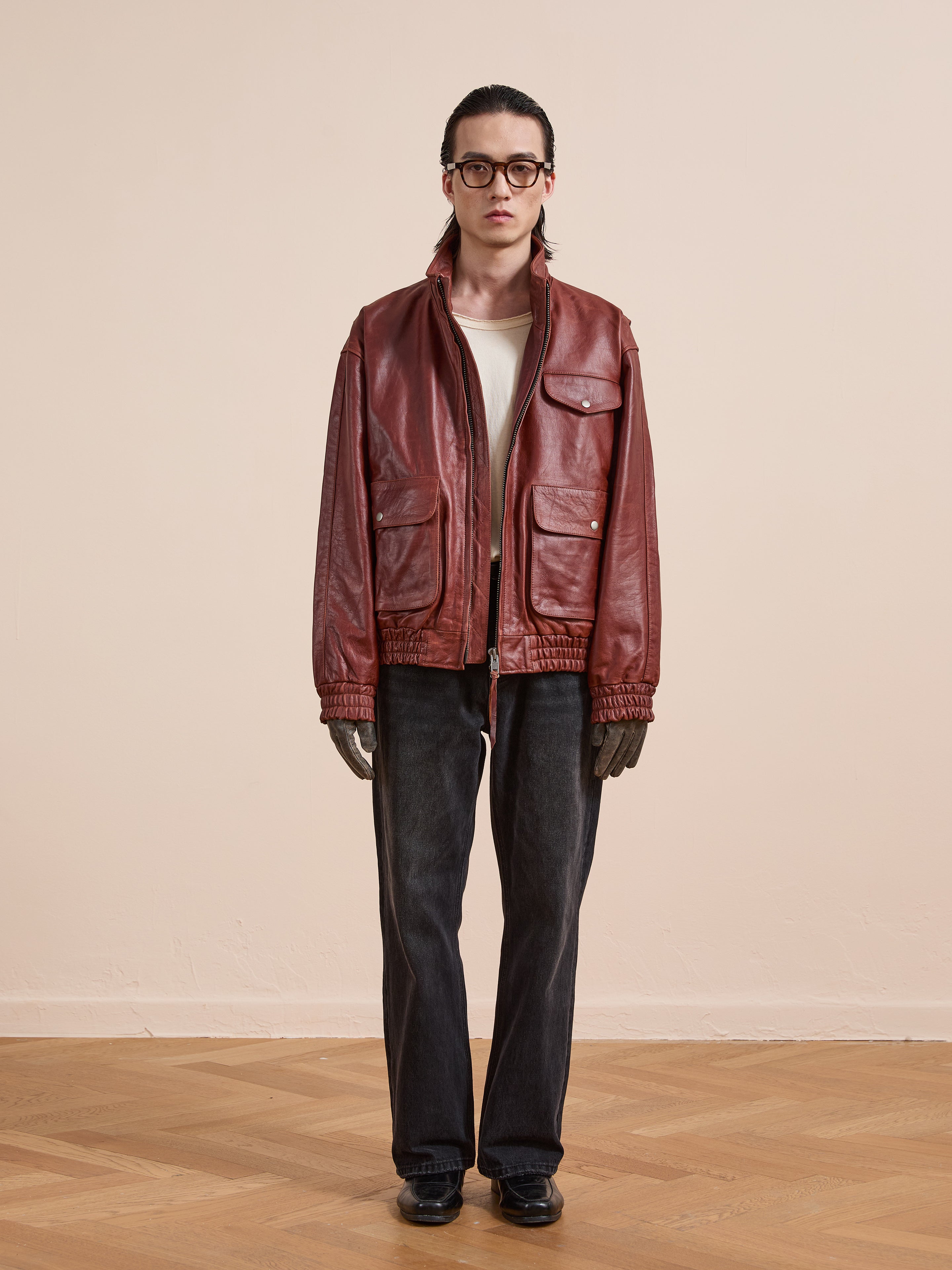 A person stands indoors facing the camera, wearing Found's Redwood Leather Bomber Jacket, a white shirt, black jeans, black shoes, dark gloves, and round glasses against a plain light-colored wall.