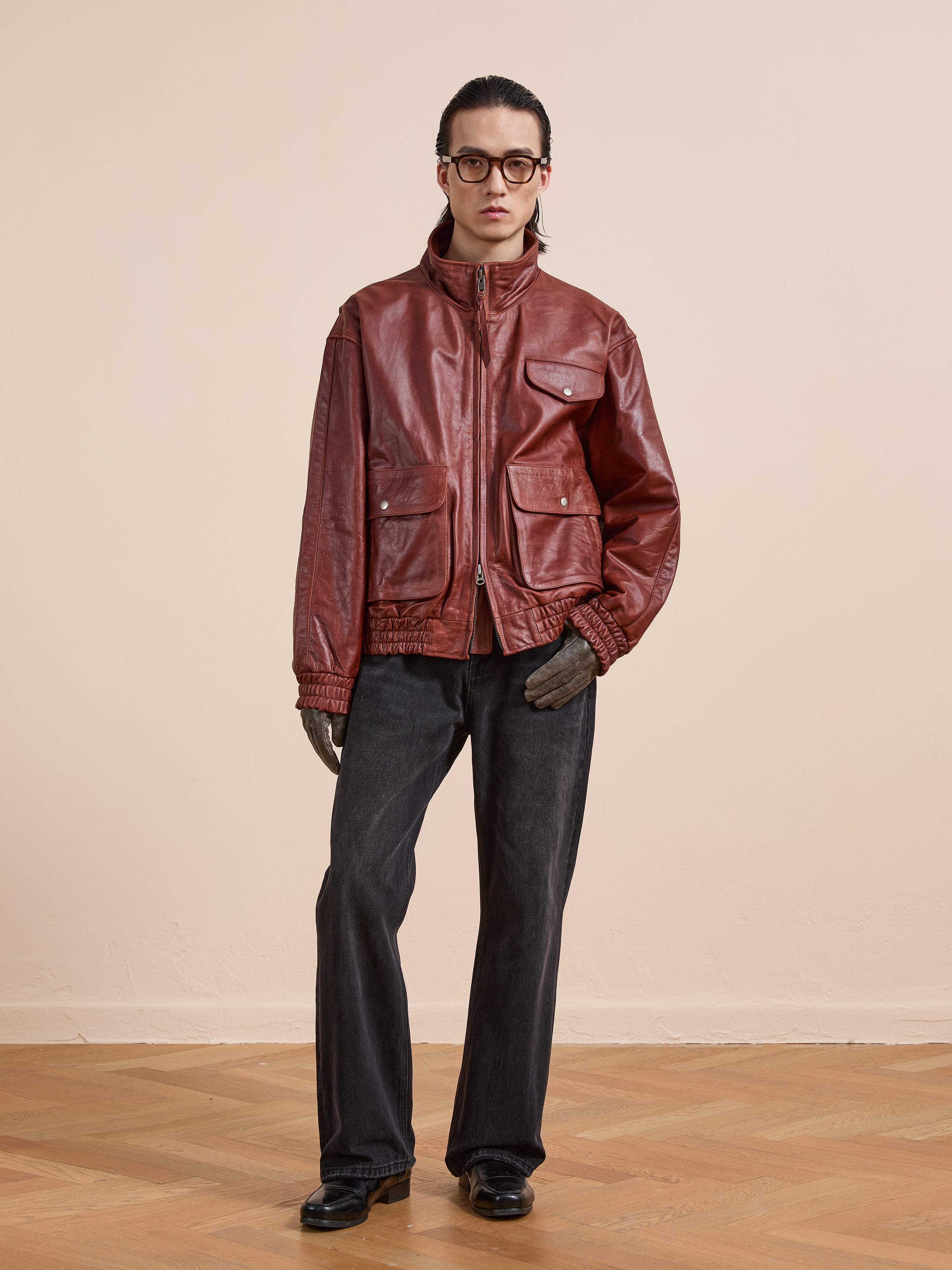 A person stands indoors facing the camera against a beige wall, wearing the Found Redwood Leather Bomber Jacket in red, paired with black pants, gloves, and glasses.