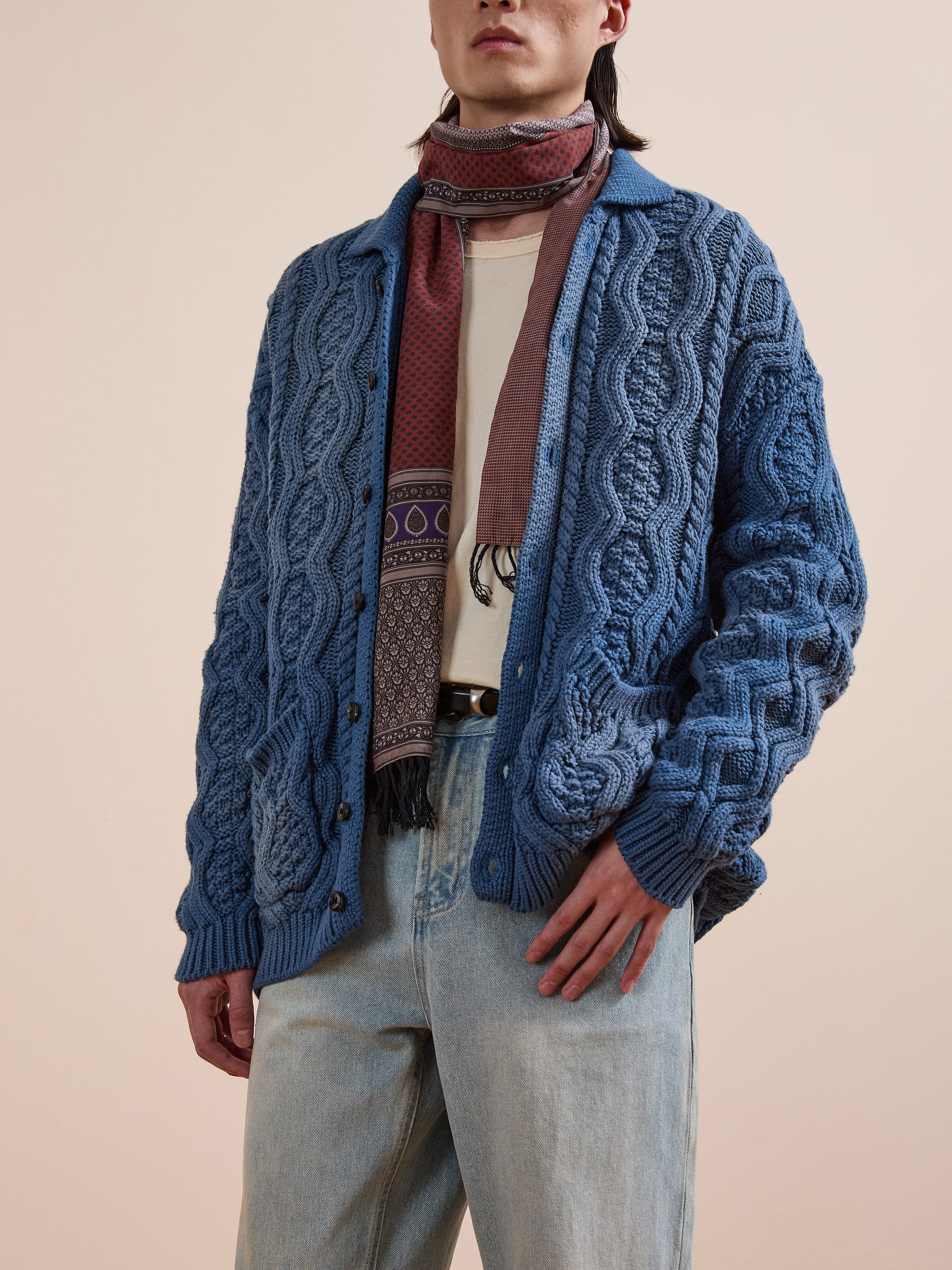 A person wears Found's blue Cable Knit Buttoned Cardigan with a multicolored scarf and light blue jeans against a plain background, giving the casual look both comfort and style.
