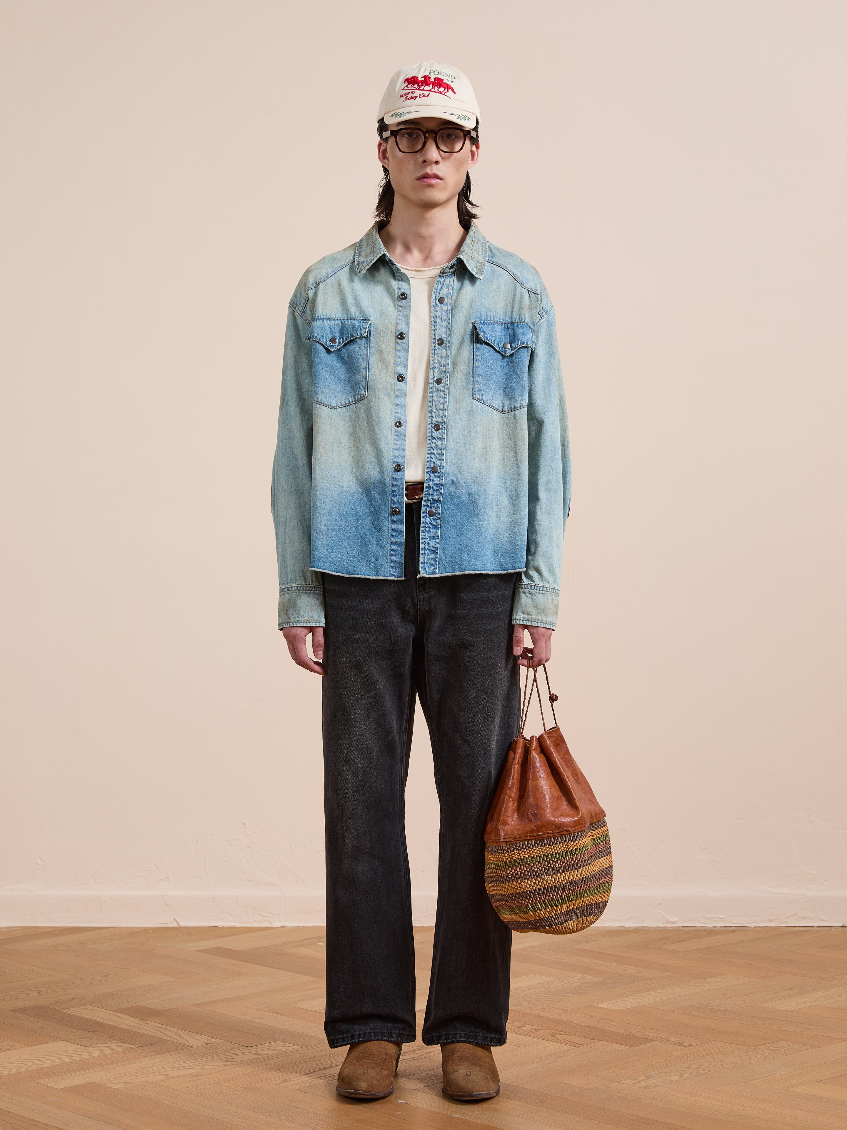 A person in a white cap, glasses, black pants, brown shoes, and the FOUND Denim Western Snap Button Shirt stands holding a brown striped bag against a plain background.