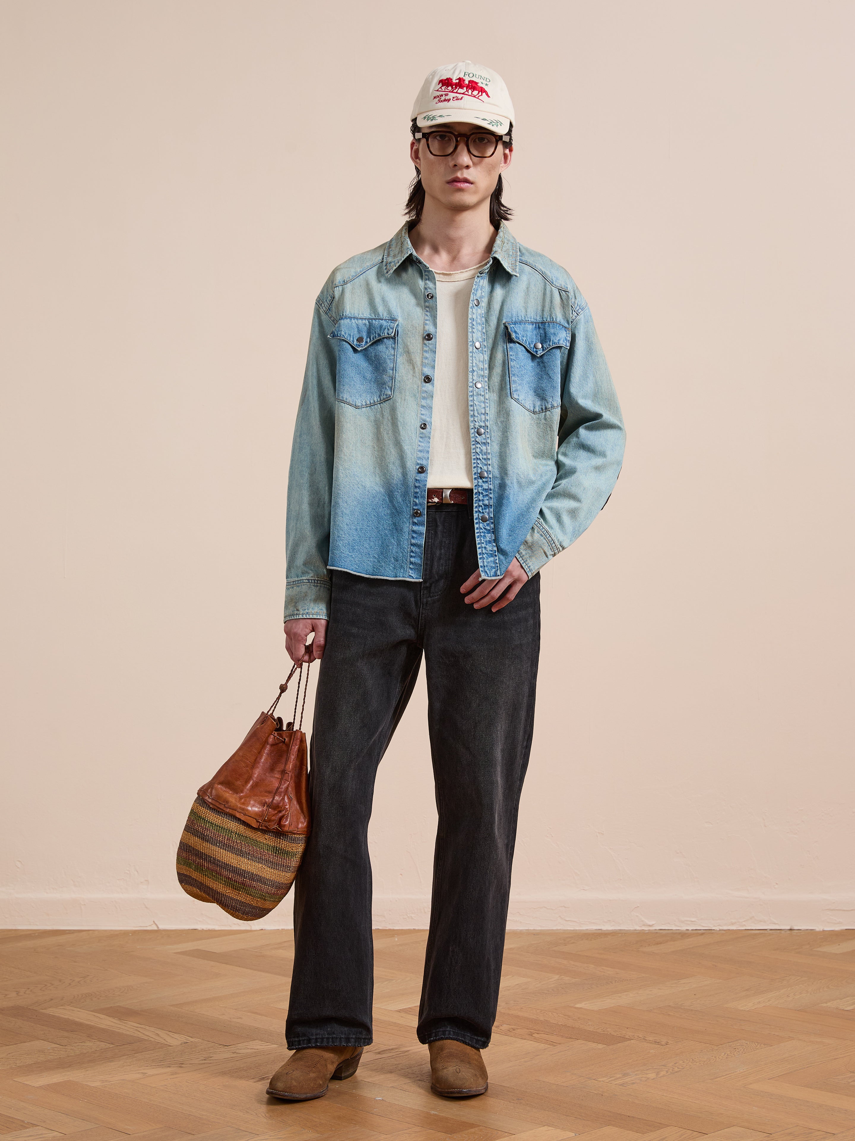 Indoors, a person wears the FOUND Denim Western Snap Button Shirt with western yoke detailing over a white t-shirt, paired with black jeans, brown shoes, glasses, a white cap, and holding a brown bag.