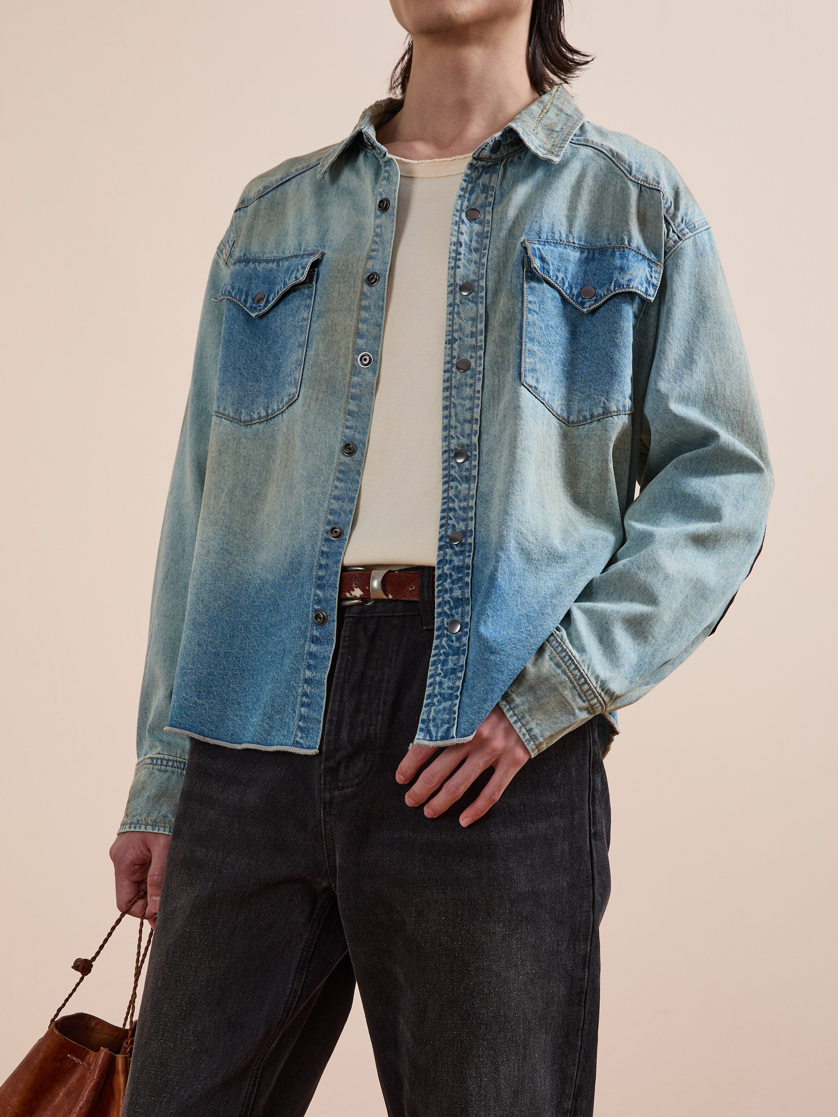 A person wearing the FOUND Denim Western Snap Button Shirt over a white tee, paired with black jeans and carrying a brown bag.