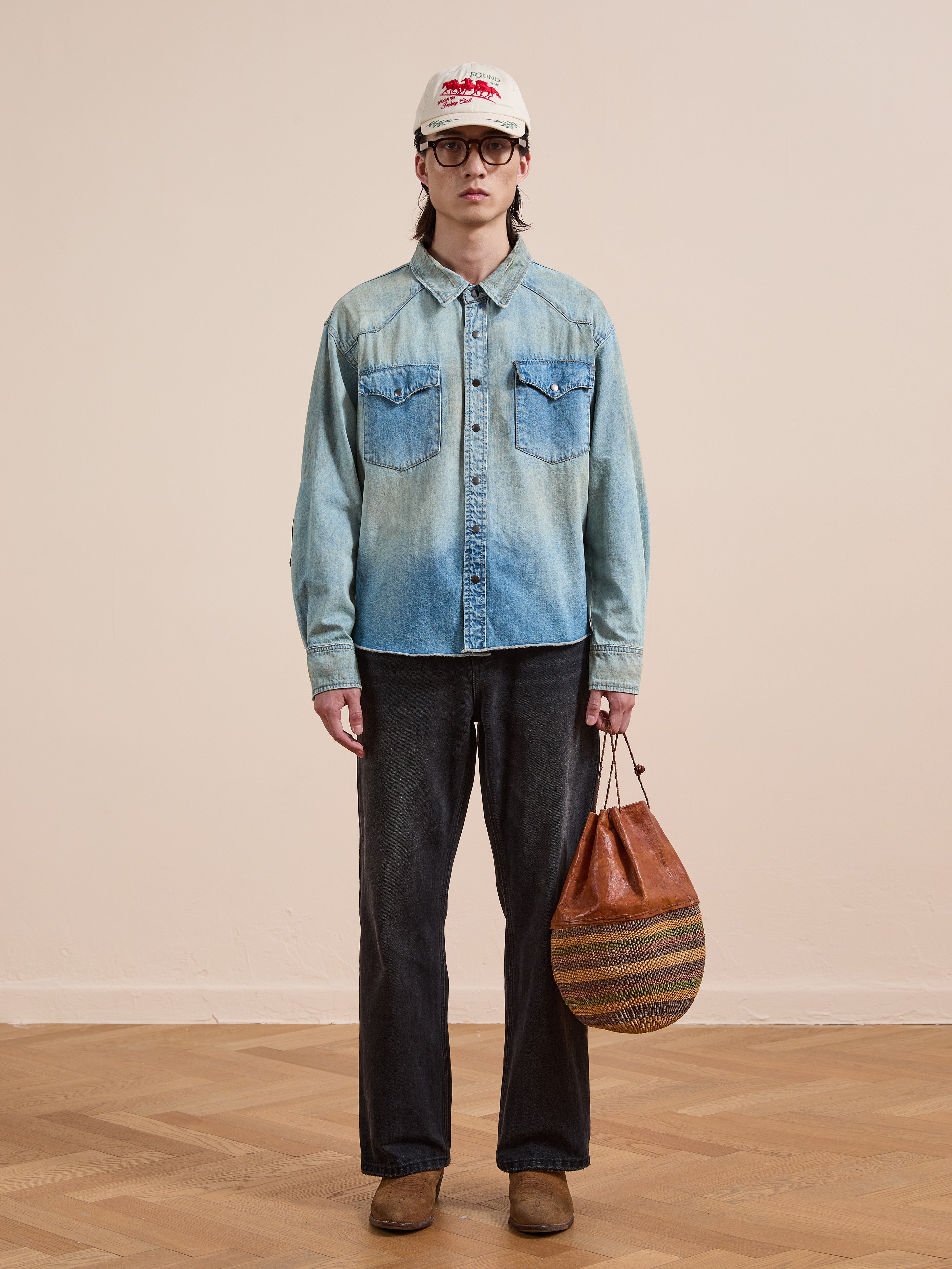 Indoors, a person wears FOUND’s Denim Western Snap Button Shirt with vintage wash and yoke detail, paired with black jeans, brown shoes, a white cap, glasses, and holds a round striped brown bag.
