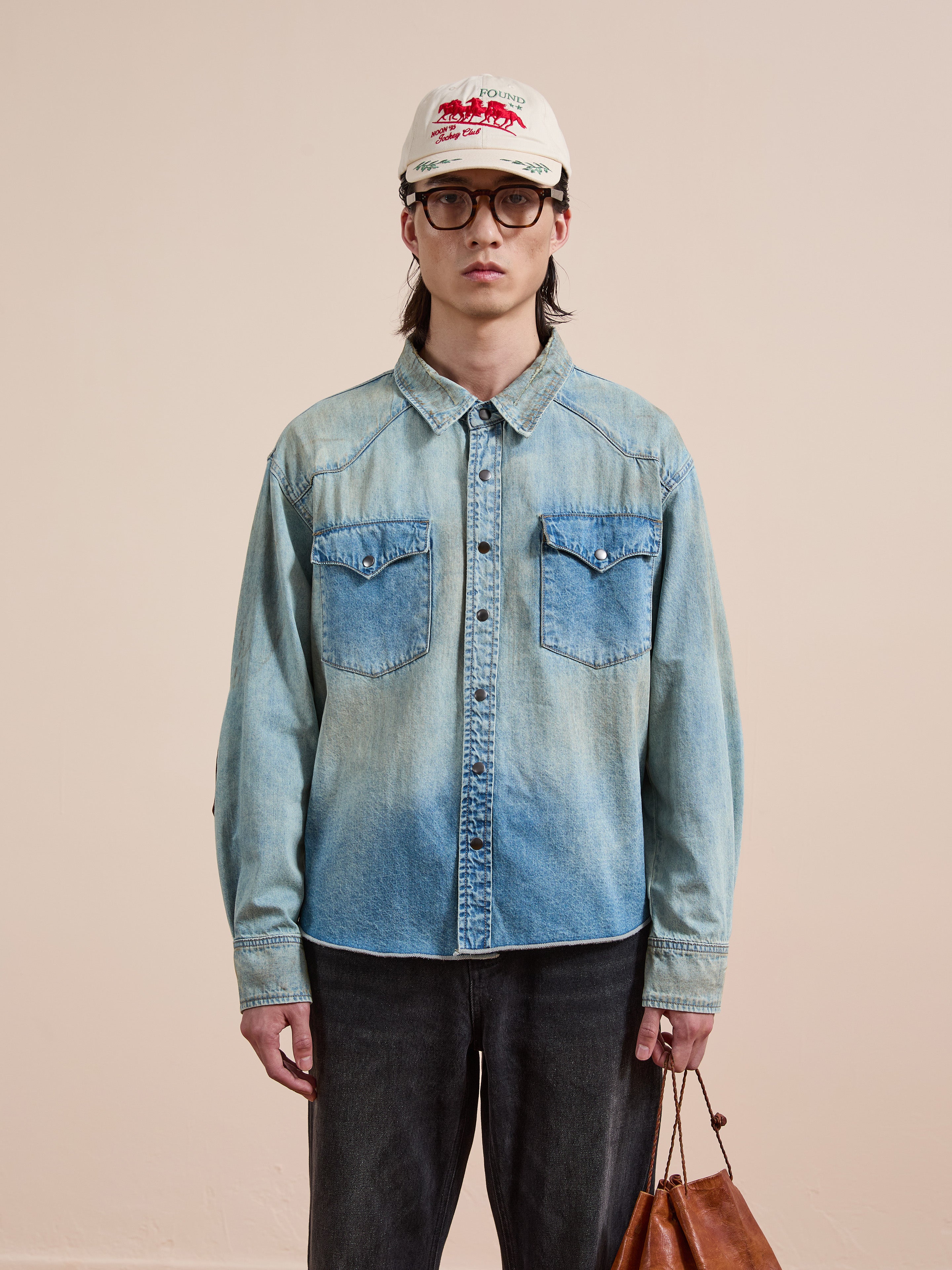 Wearing the FOUND Denim Western Snap Button Shirt with black jeans, glasses, and a white cap, a person stands against a plain background holding a brown bag.