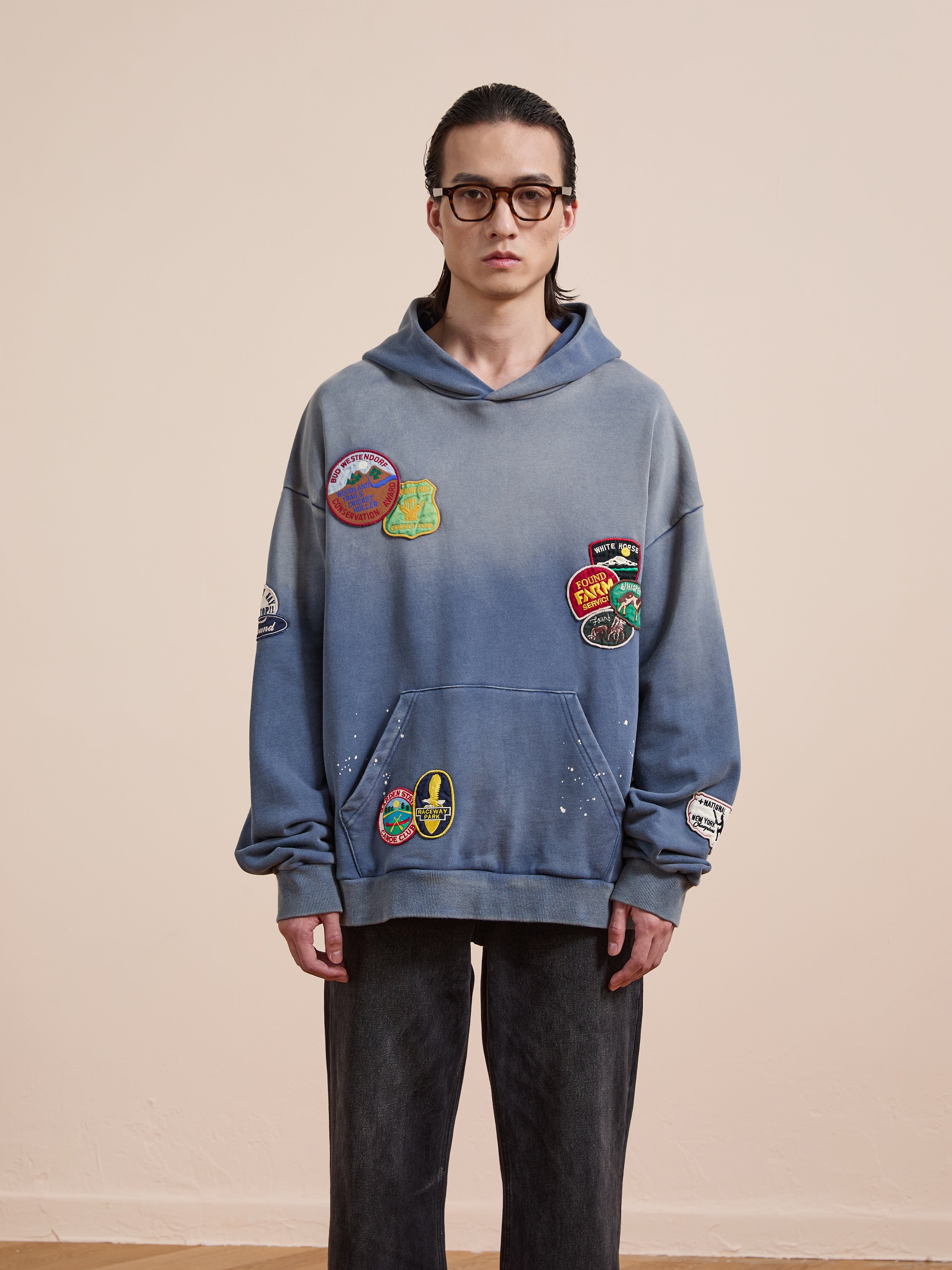 Someone in glasses models the FOUND Distressed Patch Hoodie, featuring multiple colorful patches, against a plain beige background.