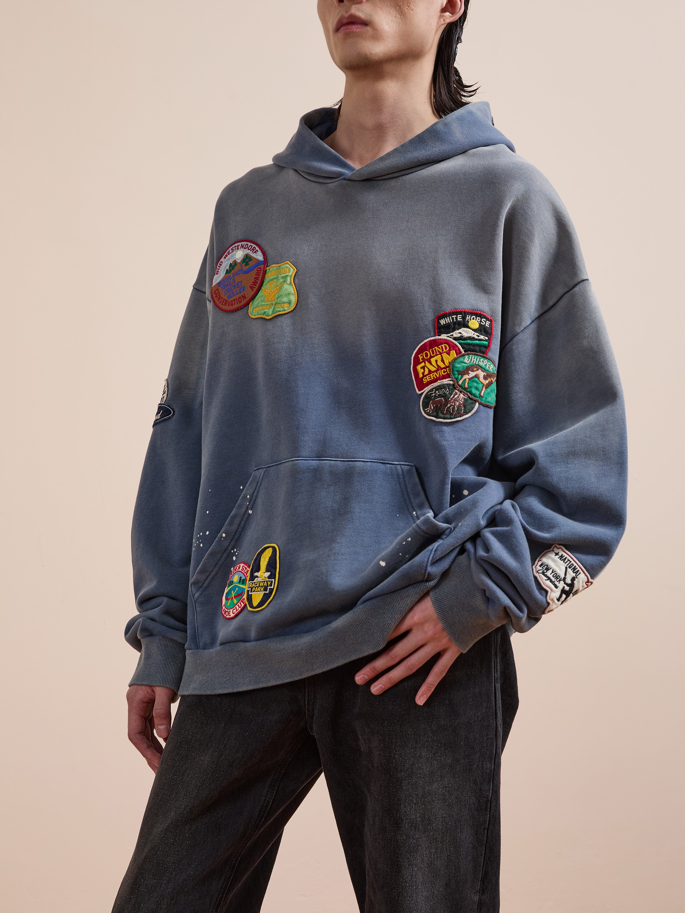 A person wearing the FOUND Distressed Patch Hoodie—a blue French Terry cotton design with colorful patches—paired with dark jeans, stands against a plain beige background.